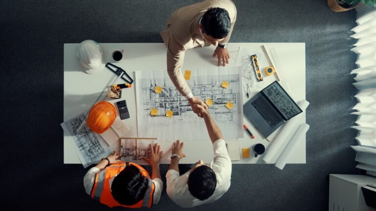 Two men at a desk shaking hands over architectural plans with a third person pointing at the plans, surrounded by tools, a laptop, and safety helmets.