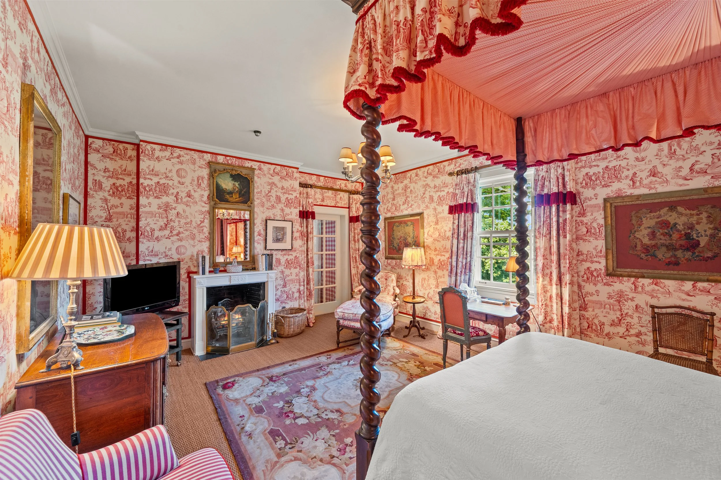 A bedroom with pink toile wallpaper, a four-poster bed with ruffled canopy, floral rug, vintage furniture, a TV, fireplace, and windows letting in natural light.