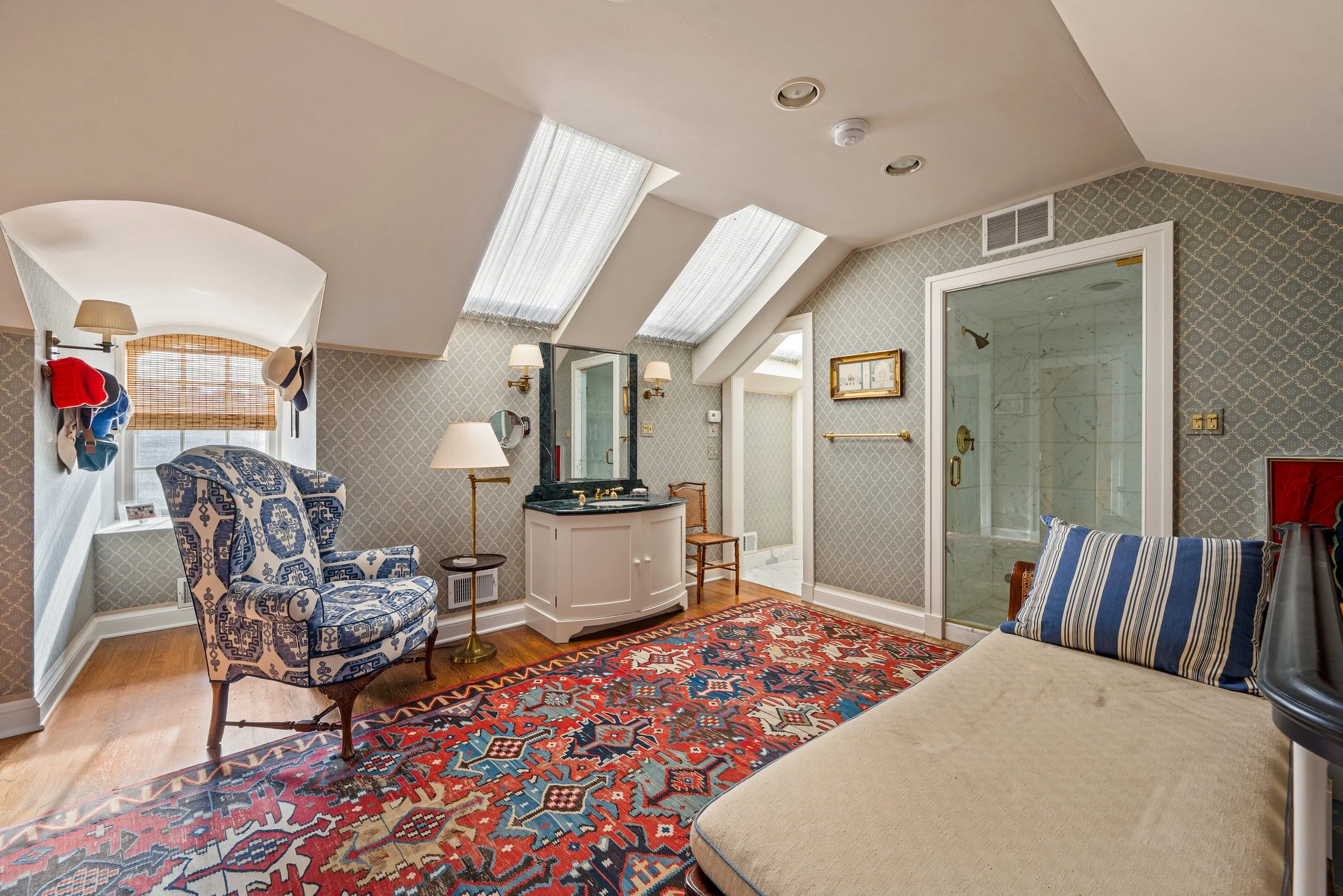 A cozy attic bedroom with a sloped ceiling and skylights, featuring a patterned armchair, a wooden chair, a side table with a lamp, a colorful area rug, patterned wallpaper, a window with blinds, a bench with a striped pillow, and a glass-enclosed sh