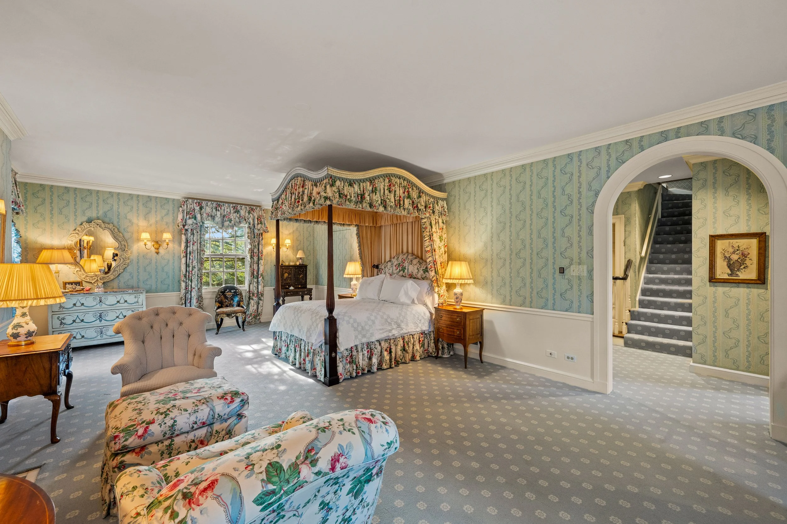 Elegant bedroom with floral decor, four-poster canopy bed, floral armchair, chaise lounge, and antique furniture.