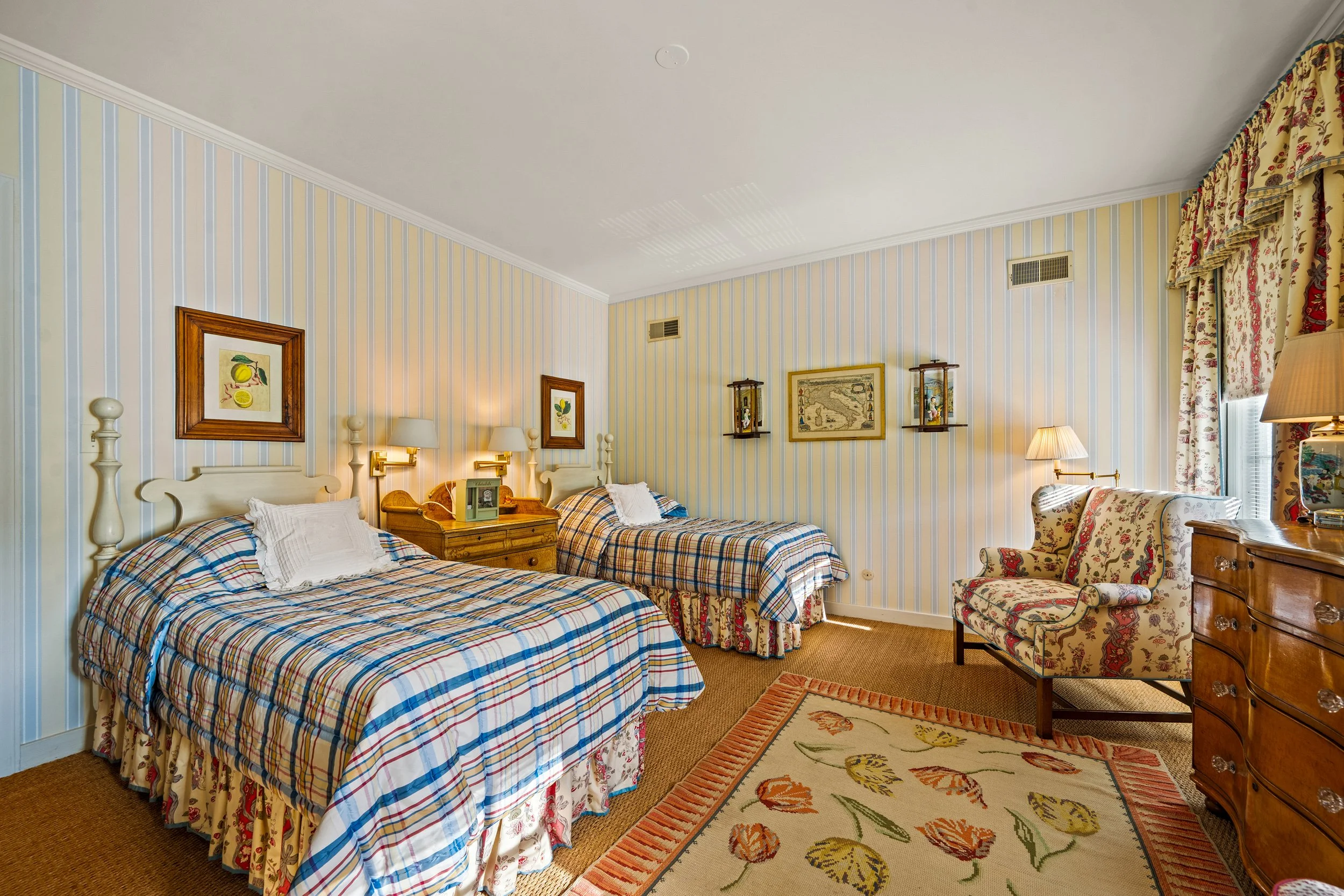 A vintage-style hotel room with two twin beds covered in plaid bedspreads, floral curtains, framed artwork on pastel-striped wallpaper, a wooden dresser with a lamp, a floral armchair, and an area rug with tulip patterns.
