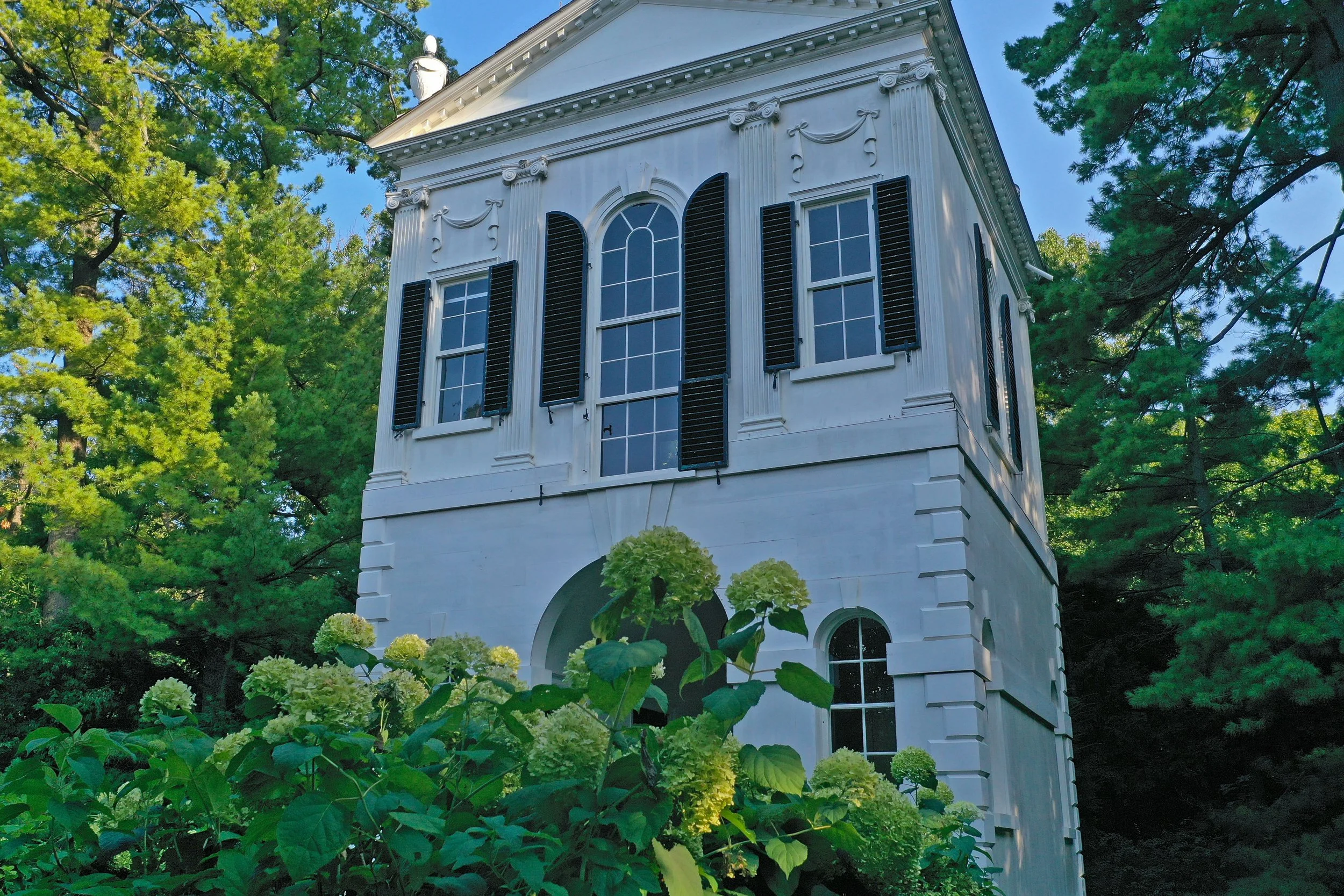 A white, two-story historic building with black shutters and arched windows, surrounded by green trees and flowering bushes.
