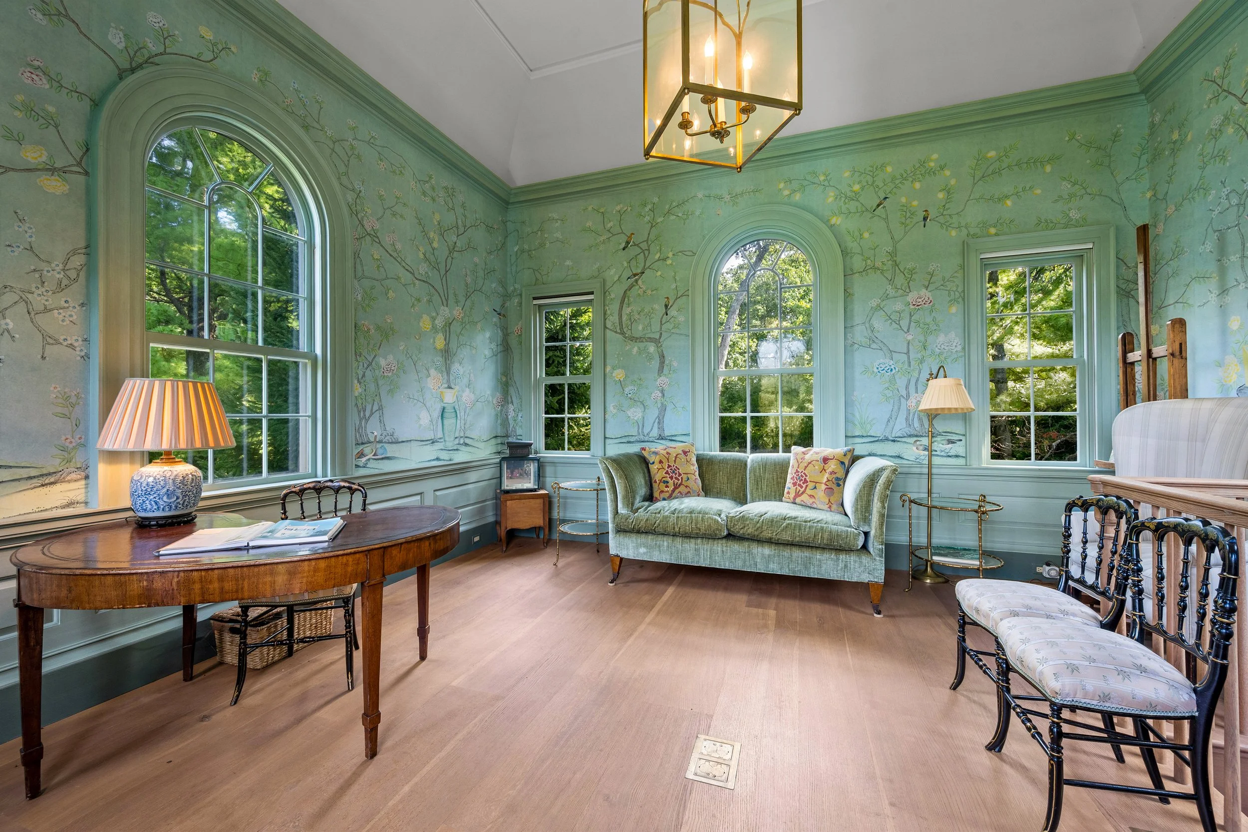 A vintage-style living room with green walls featuring floral murals, large arched windows with views of trees outside, a green velvet sofa with decorative pillows, a wooden table with a lamp, and a combination of wooden and upholstered chairs, illum