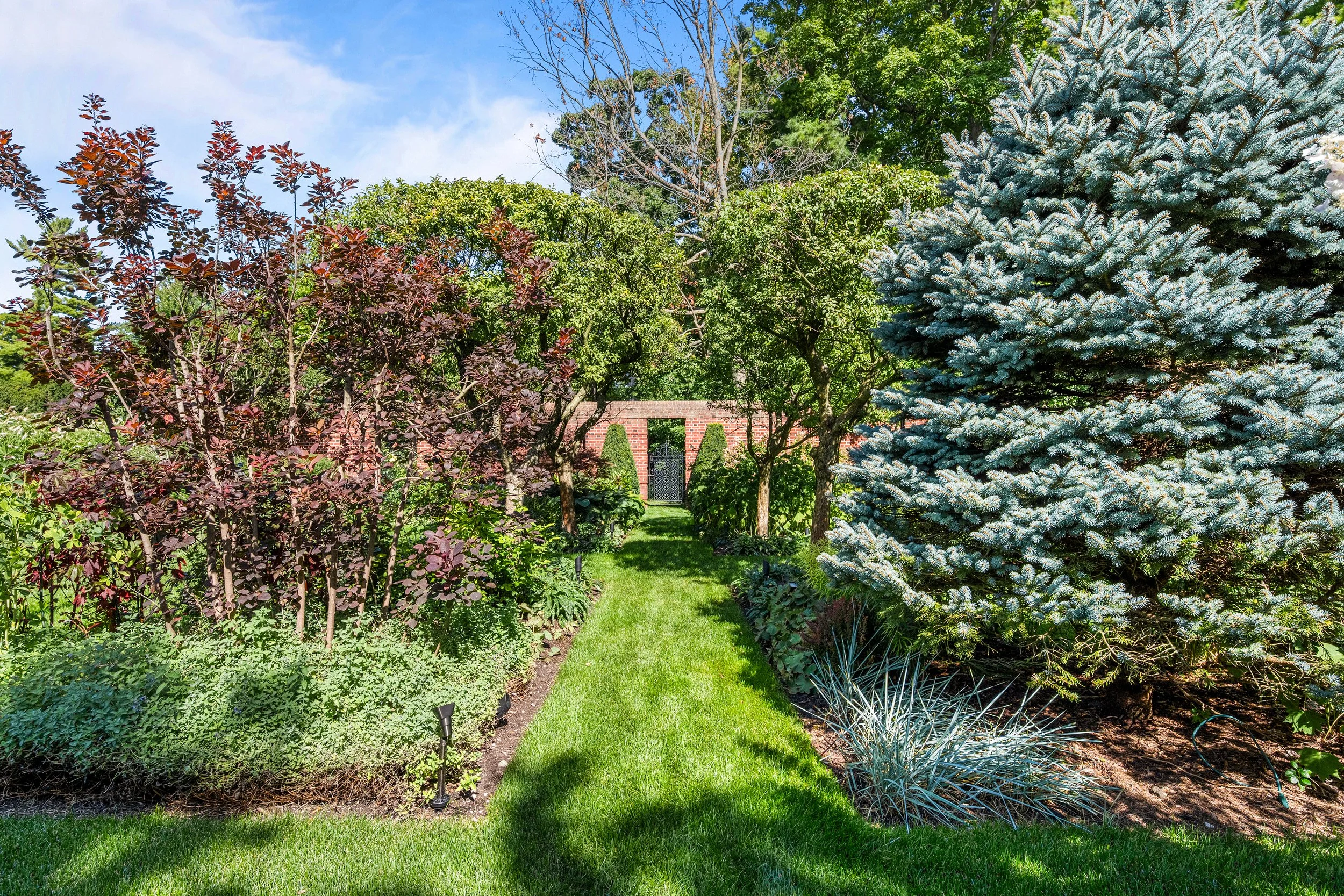 A lush garden with a narrow grassy pathway bordered by various trees and shrubs, including a tall evergreen on the right, and a brick structure with an arched opening and gate at the end of the path, under a blue sky.
