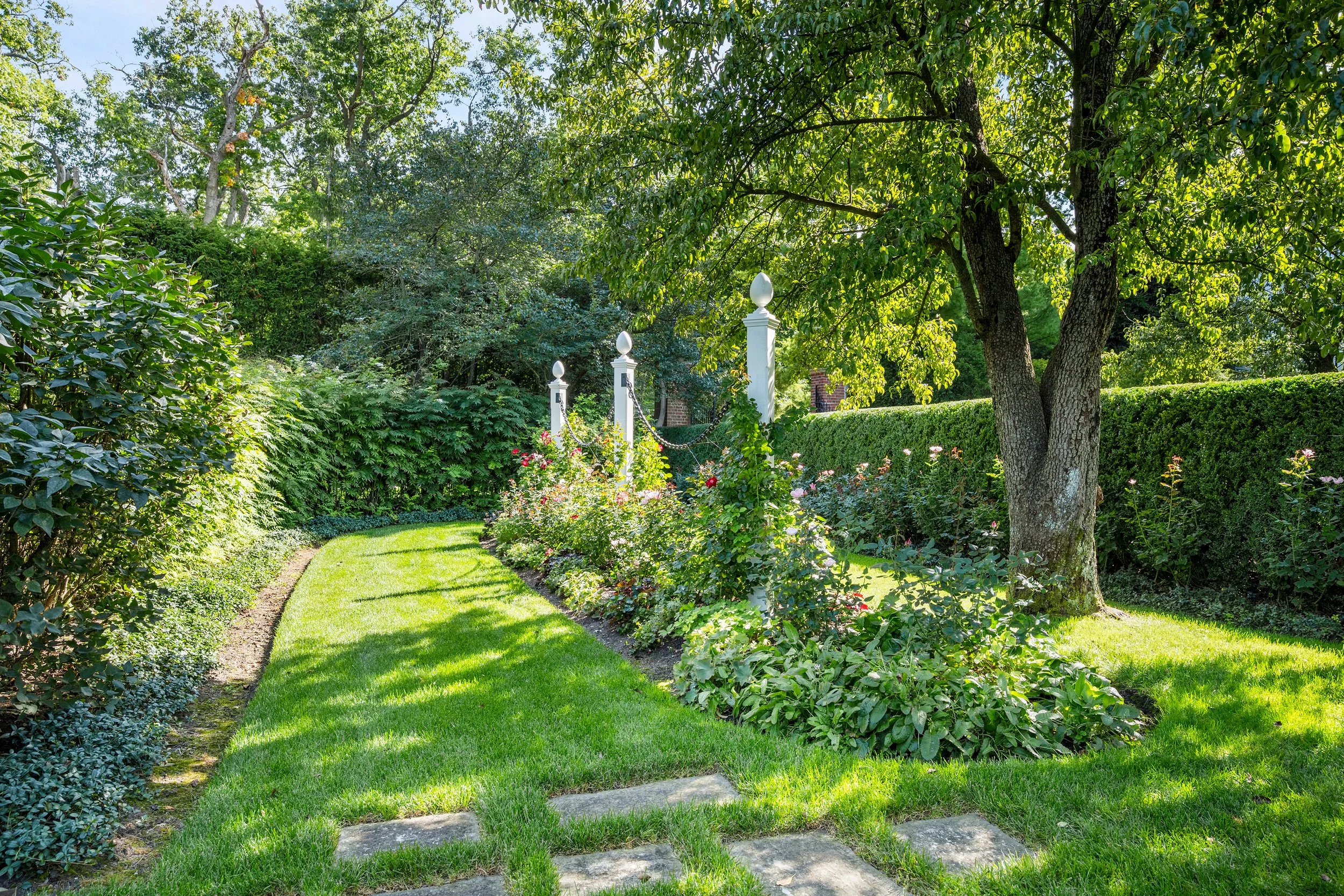 A lush garden with a manicured lawn, a flower bed with blooming pink and white roses, a large leafy tree, neatly trimmed hedges, and white decorative posts connected with chains.