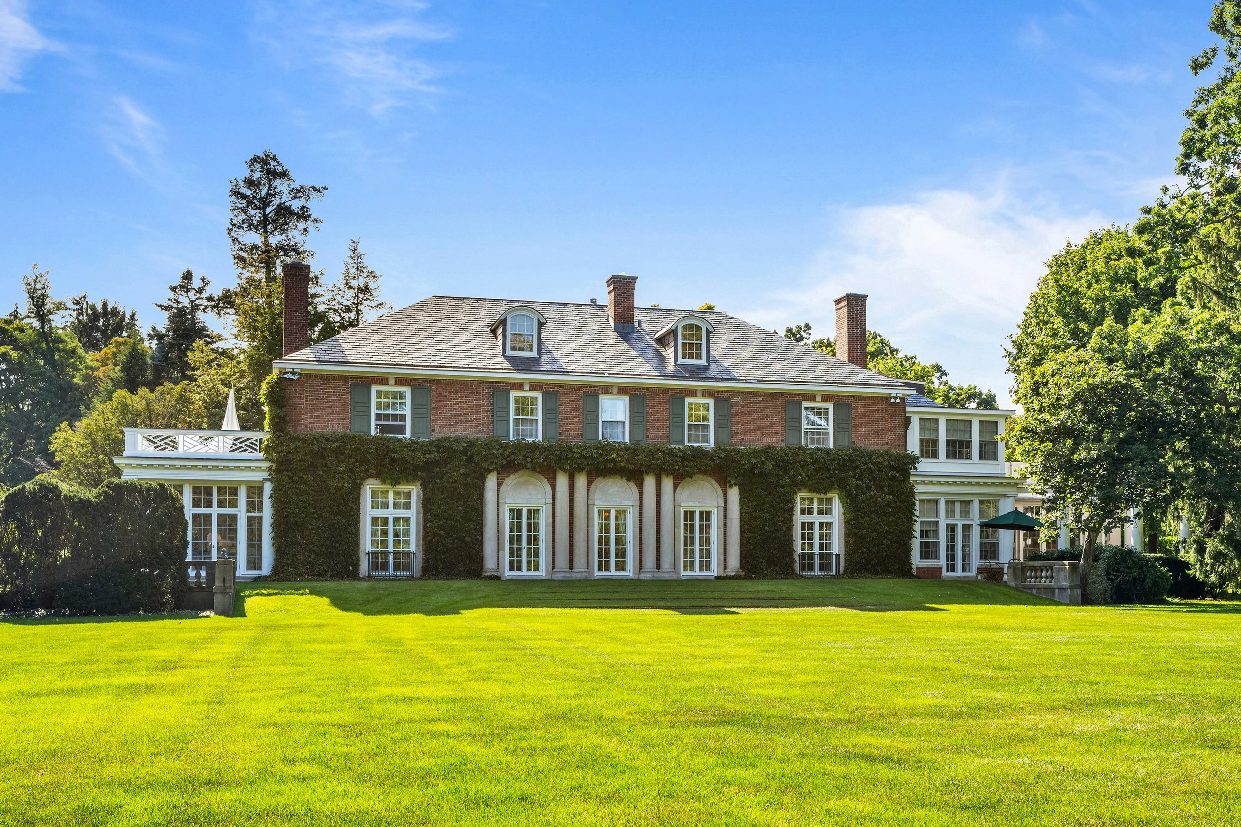 Large brick house with multiple chimneys, green shutters, and a combination of traditional and modern architecture, set on a lush green lawn with trees and a blue sky.