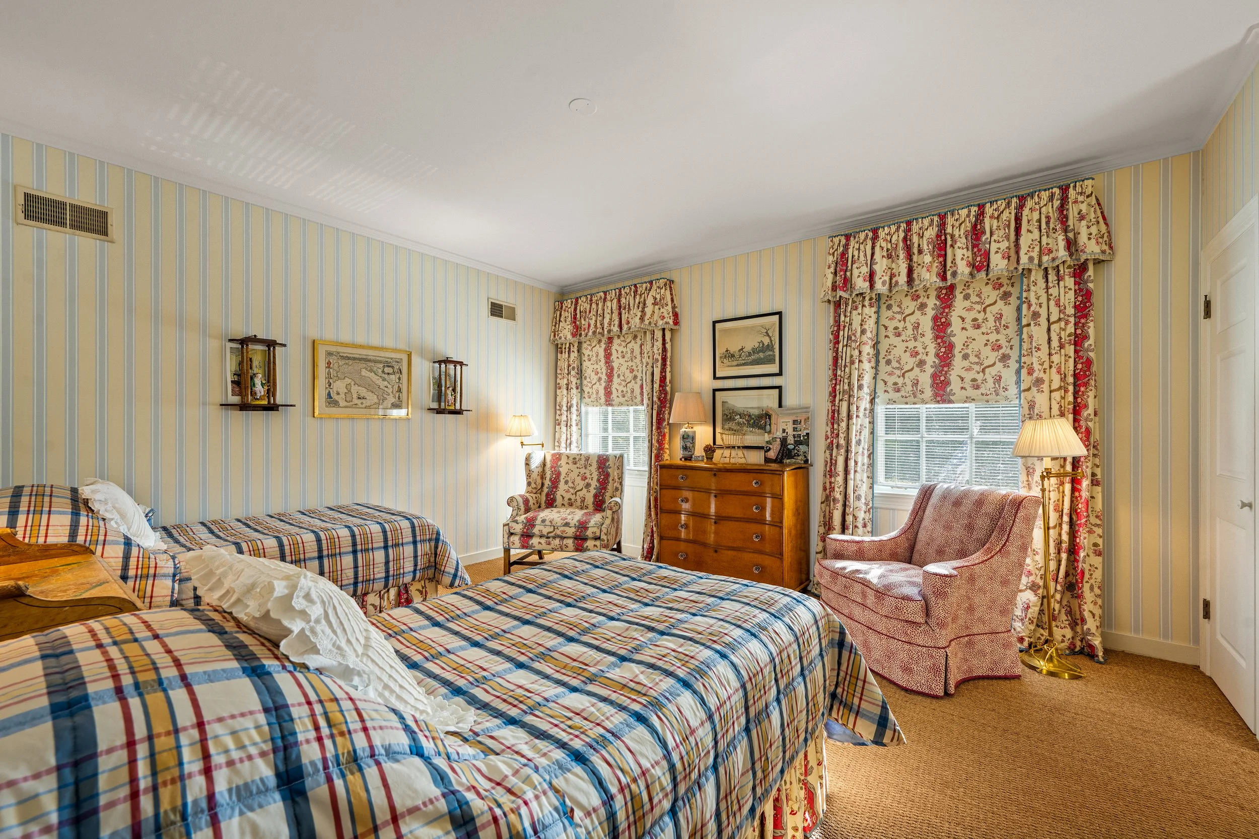 A bedroom with two beds with plaid and floral bedding, a vintage dresser with framed pictures, two armchairs with floral upholstery, patterned curtains, and striped wallpaper.
