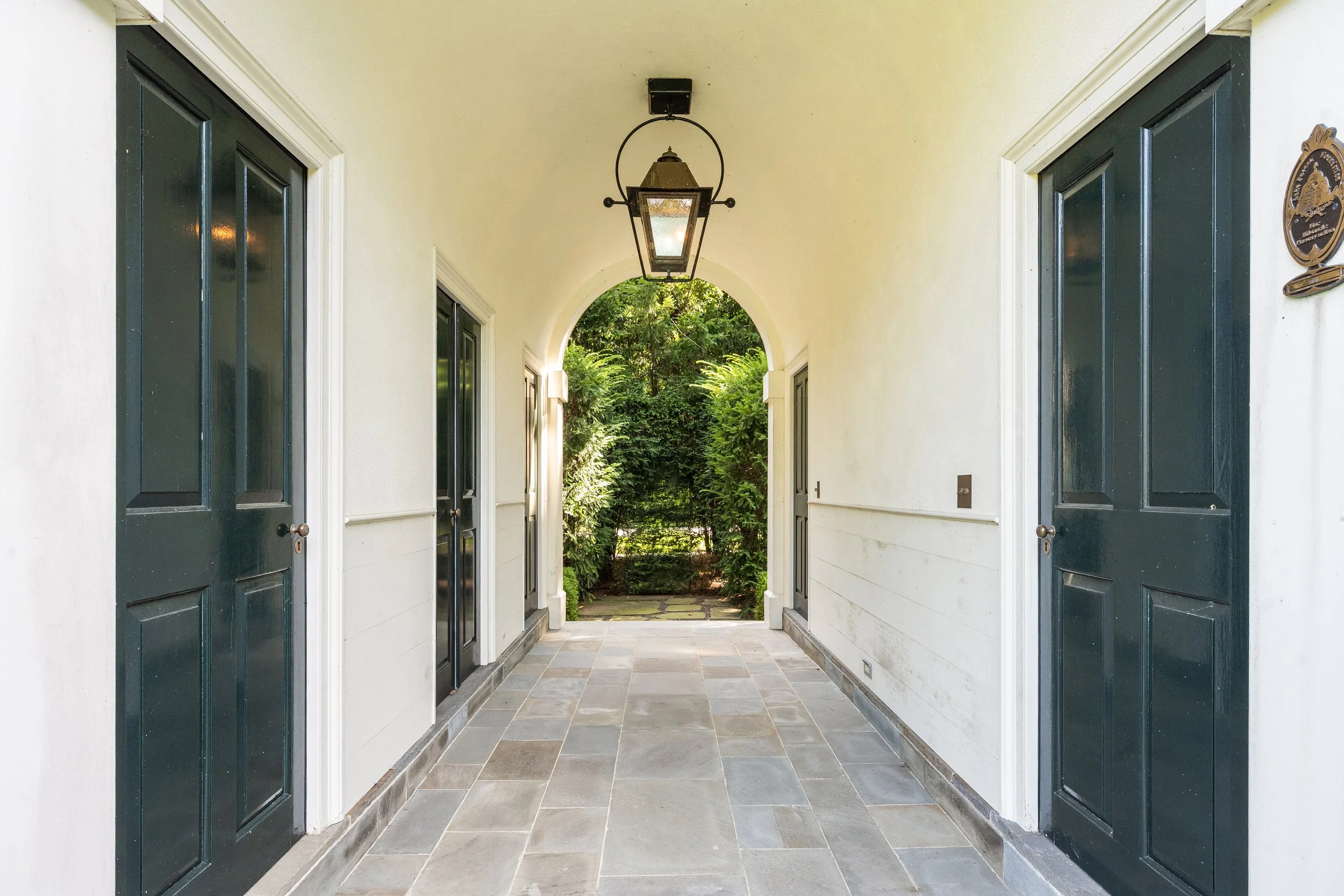 Covered walkway with two black doors on each side, a lantern hanging from the ceiling, and greenery at the end of the corridor.