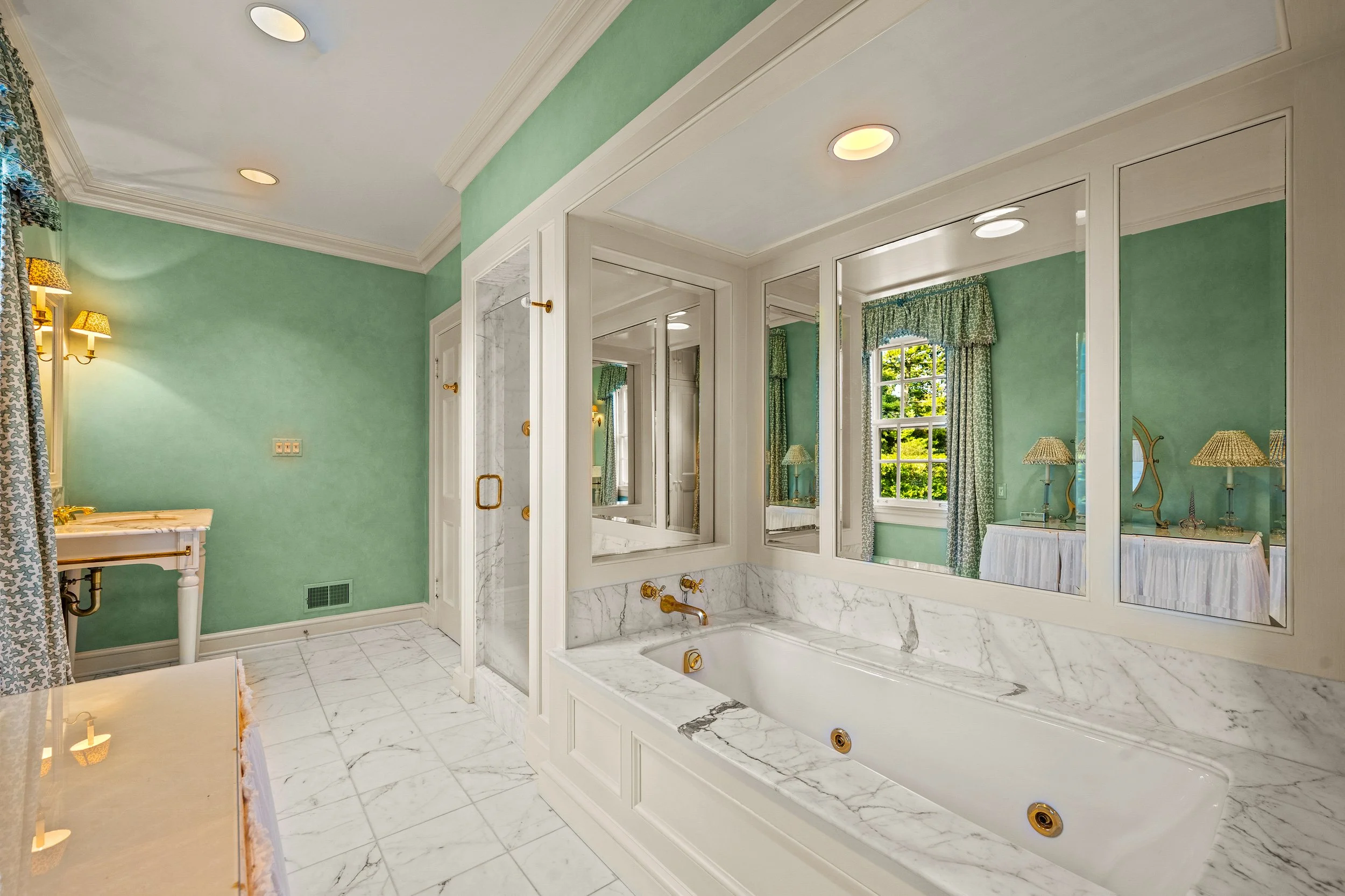 A luxurious bathroom with a marble bathtub, large mirrors, green walls, and matching curtains, with a view of a window showing greenery outside.
