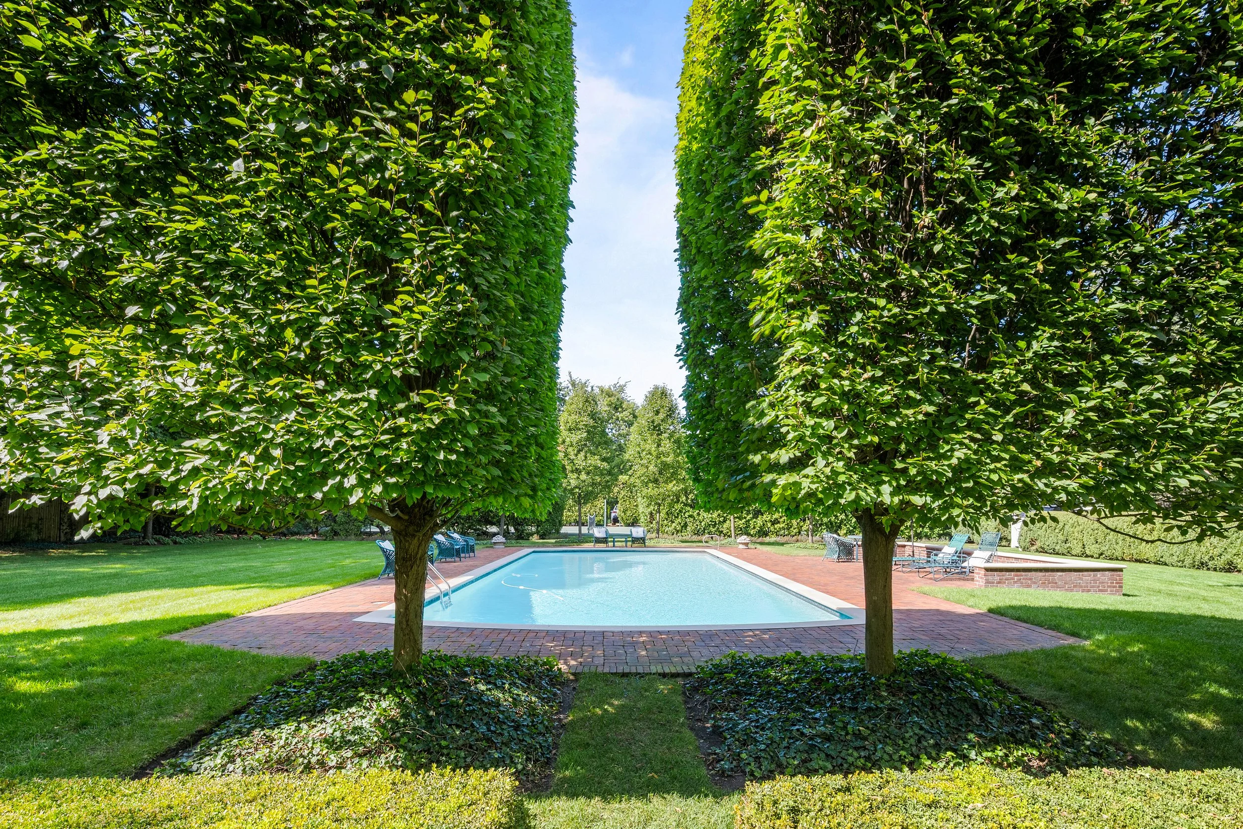 A swimming pool surrounded by green grass, with two large leafy trees framing the view and lounge chairs on a brick patio.