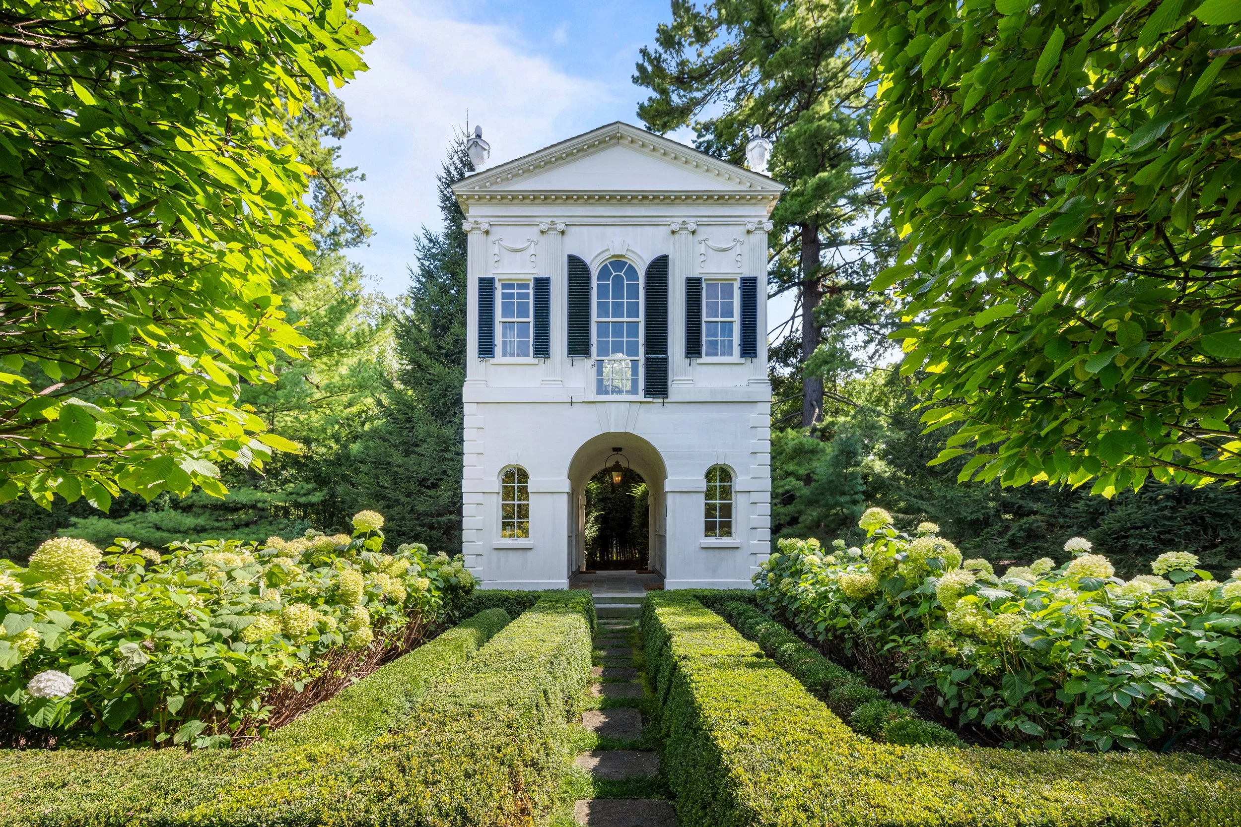 White two-story house with black shutters and arched windows, surrounded by lush green gardens and trees.