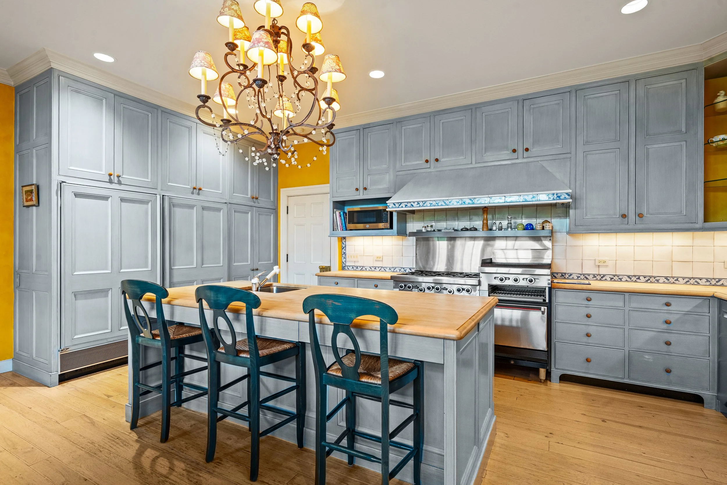 A spacious kitchen with light blue cabinets, a large wooden island with three chairs, a chandelier overhead, stainless steel appliances, and a yellow wall with a small picture frame.