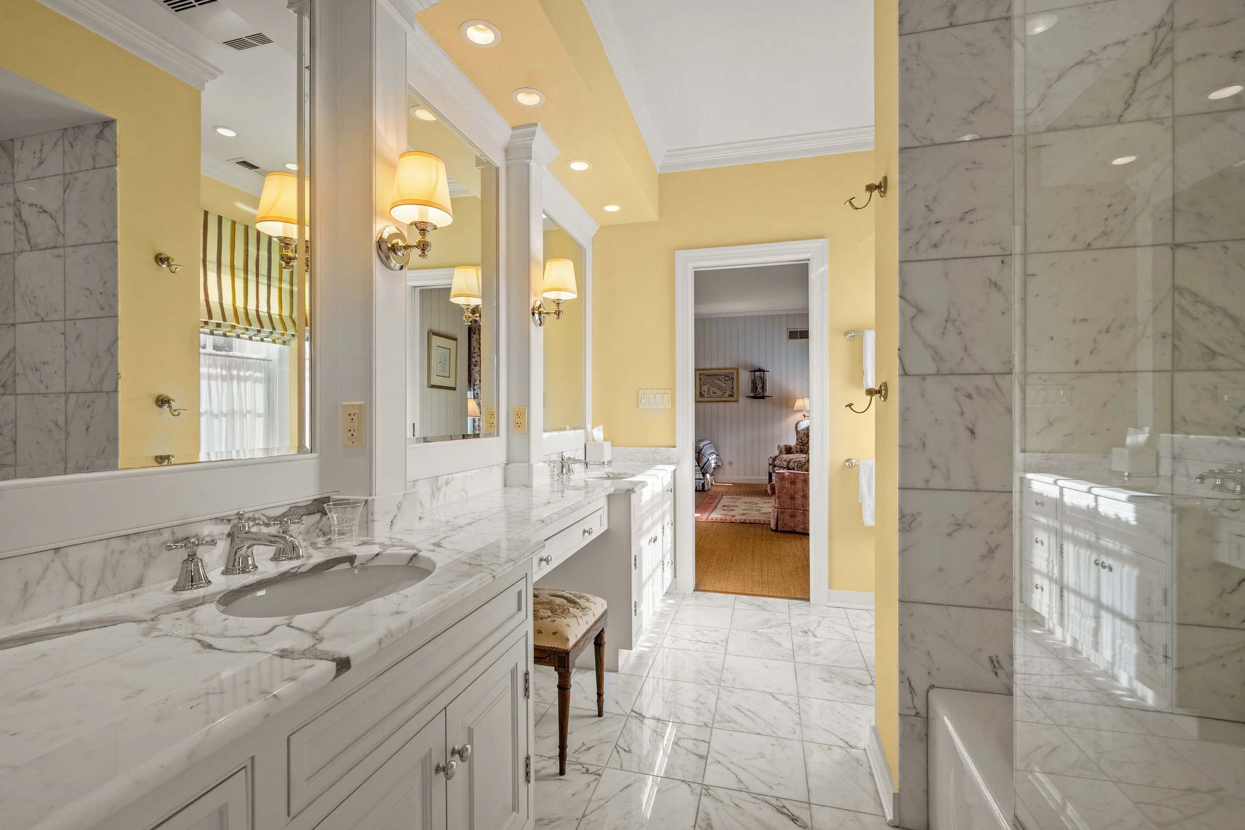 Bathroom with marble countertops, yellow walls, and mirrors with lights, leading to a bedroom with striped wallpaper and furniture.