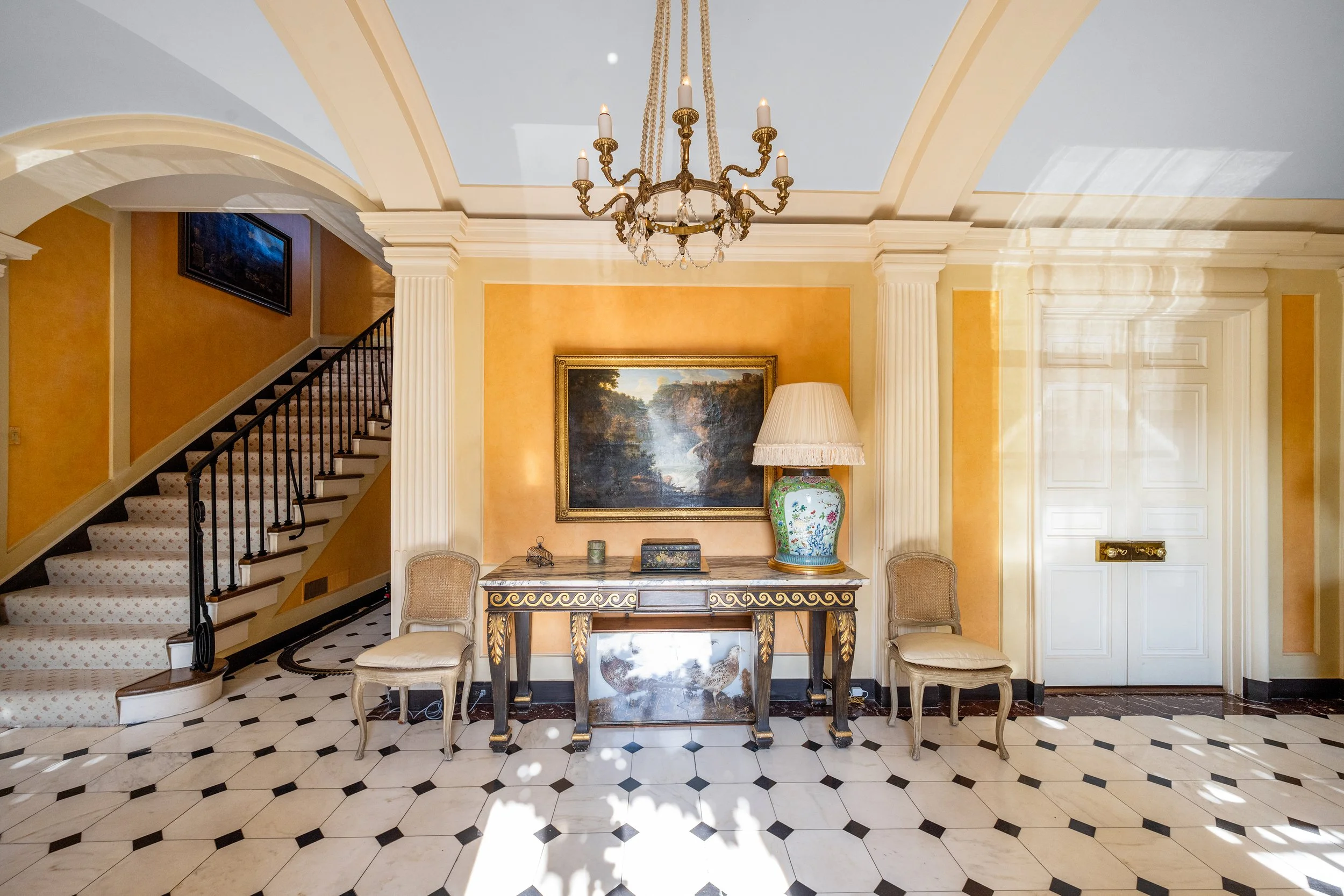 Elegant interior with a black and white checkered marble floor, yellow walls, white columns, and a luxurious chandelier. A console table with a marble top, decorative painting, a large vase with a lamp, and two antique chairs are arranged symmetrically against the wall. An staircase with a patterned carpet and a black railing rises on the left side, while a white door with gold hardware is on the right.