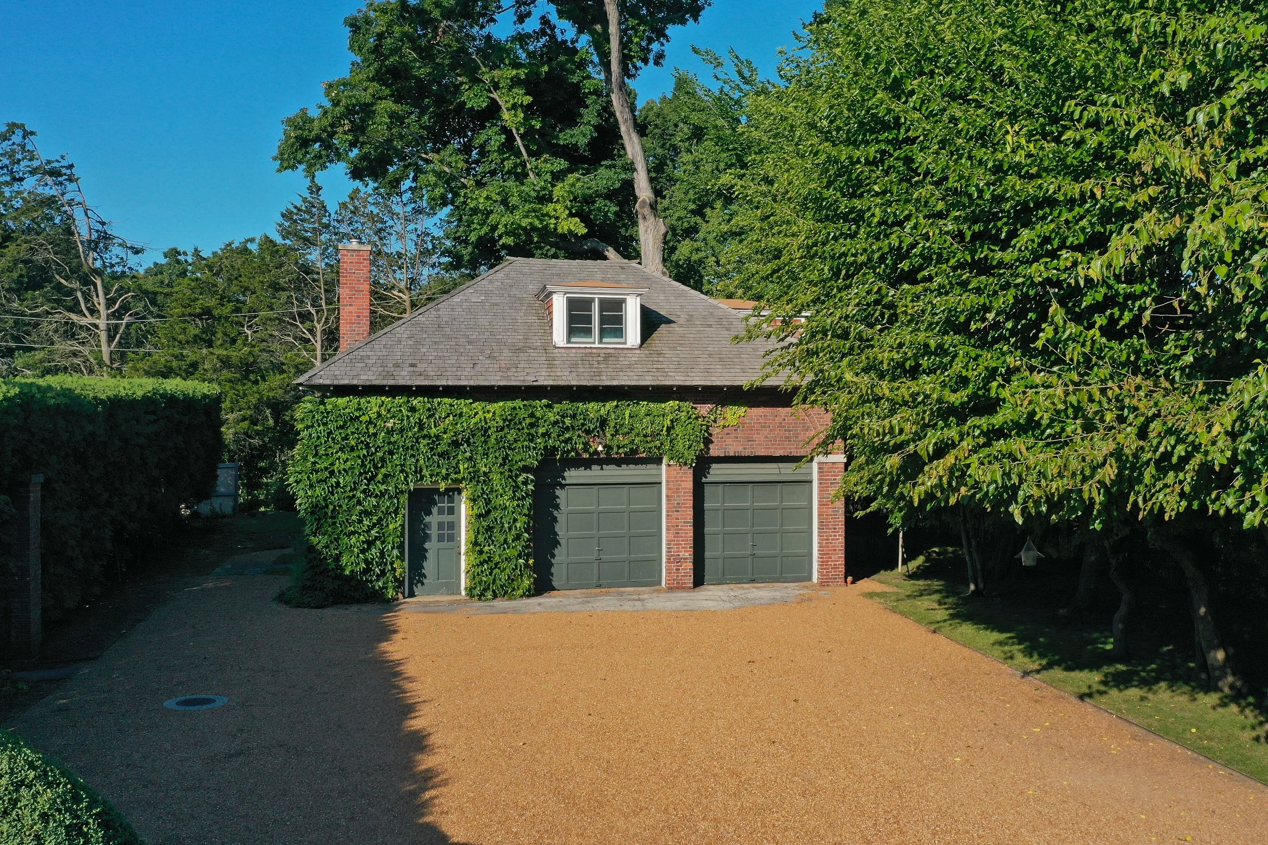 A detached garage with two green doors, brick walls, and a dormer window on the roof, surrounded by greenery and trees under a clear blue sky.