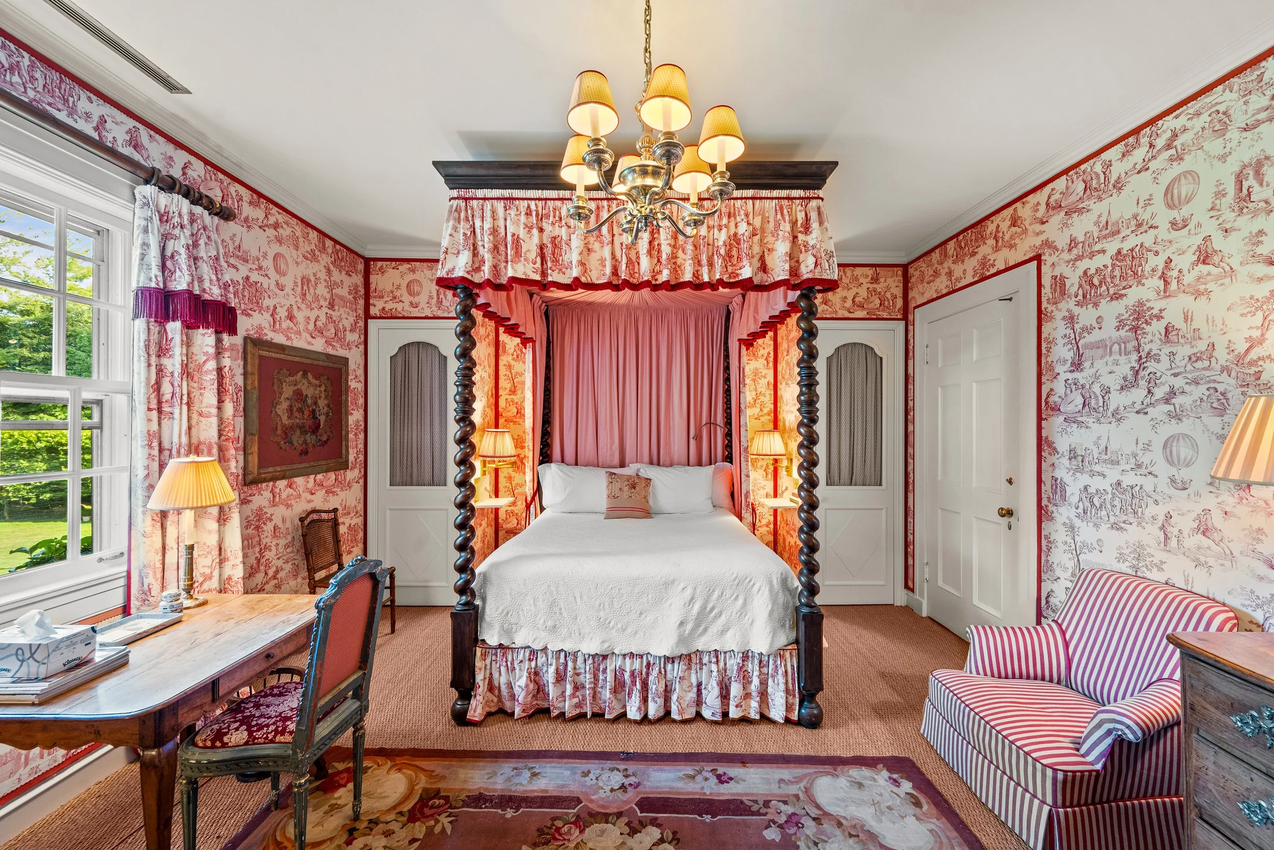 A bedroom with a decorated four-poster bed draped with pink fabric, floral wallpaper, striped armchair, wooden desk with a chair, window with floral curtains, and warm lighting from lamps and a chandelier.