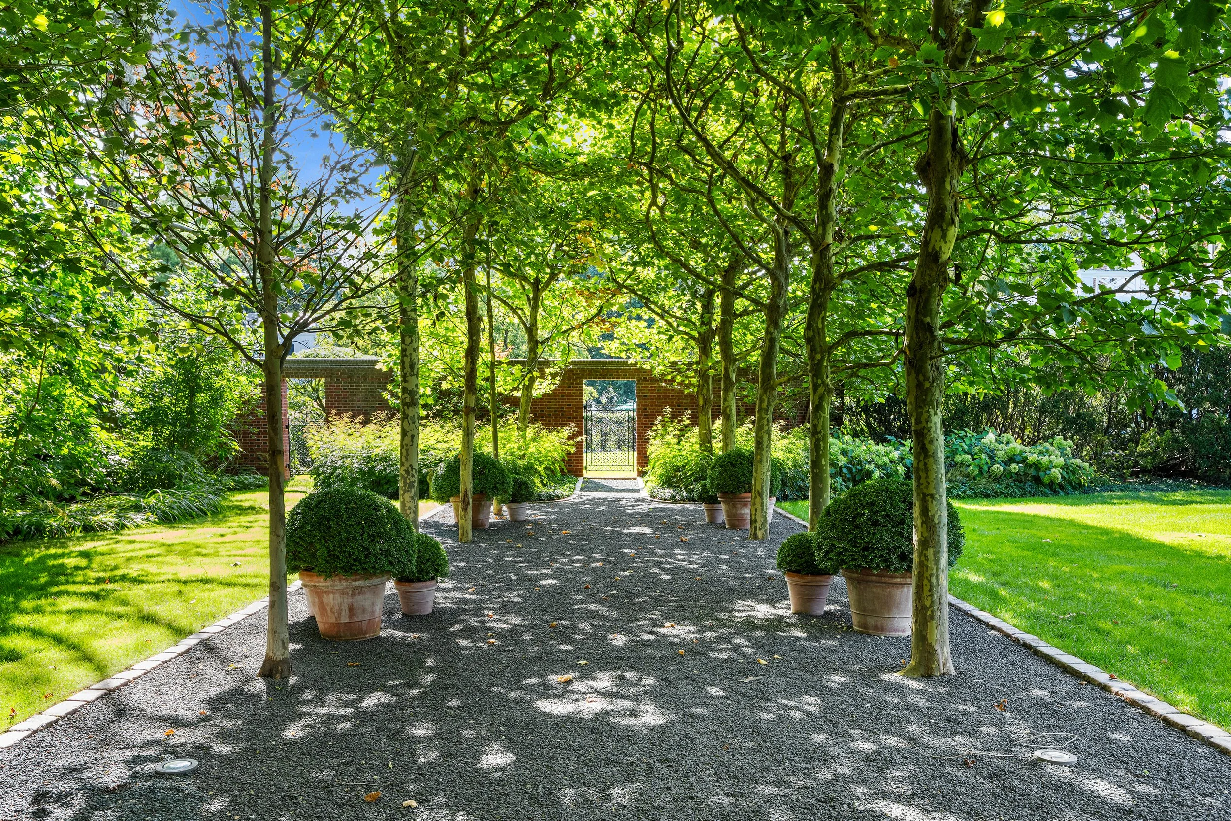 A shaded gravel pathway lined with potted green bushes under a canopy of trees, leading to a brick wall with an arched gate in a lush garden.