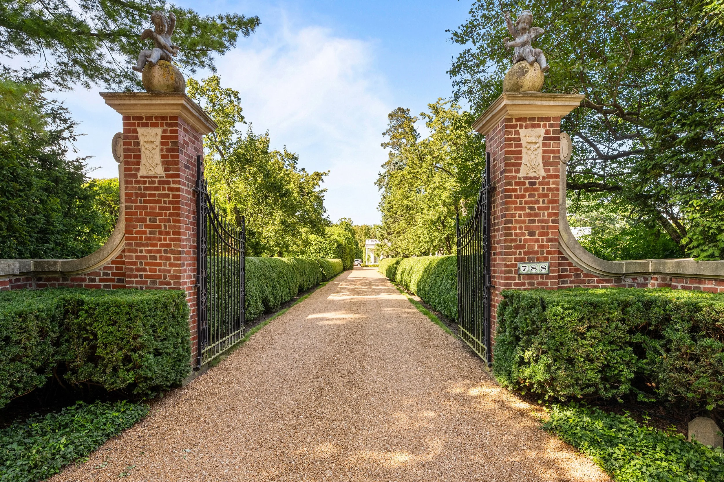 Entrance gate with brick pillars, black wrought iron gates, and surrounding green shrubs leading onto a pathway lined with trees, under a partly cloudy sky.
