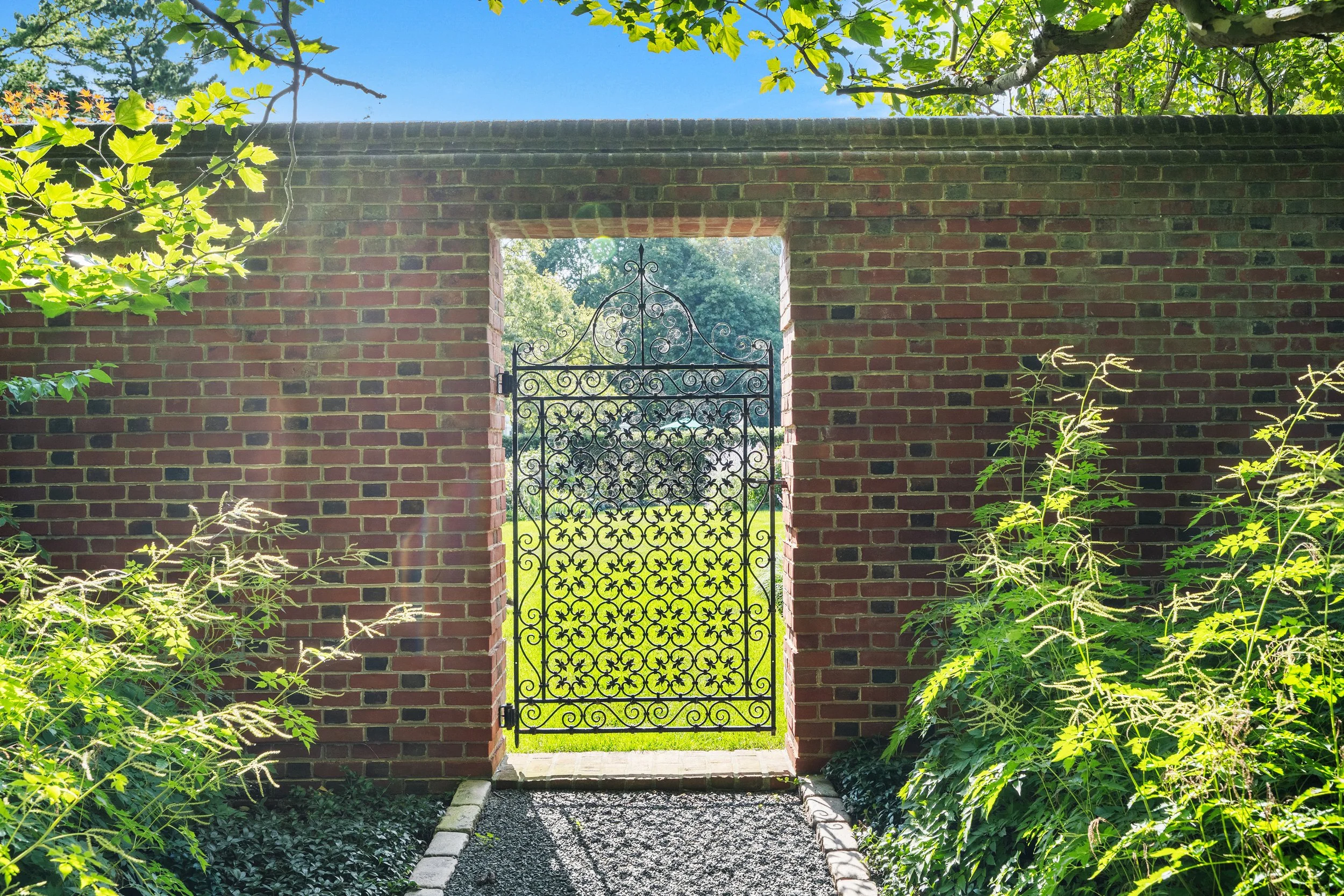 A black wrought iron gate in a brick wall, opening to a lush green yard with trees and grass, under a clear blue sky with sunlight.