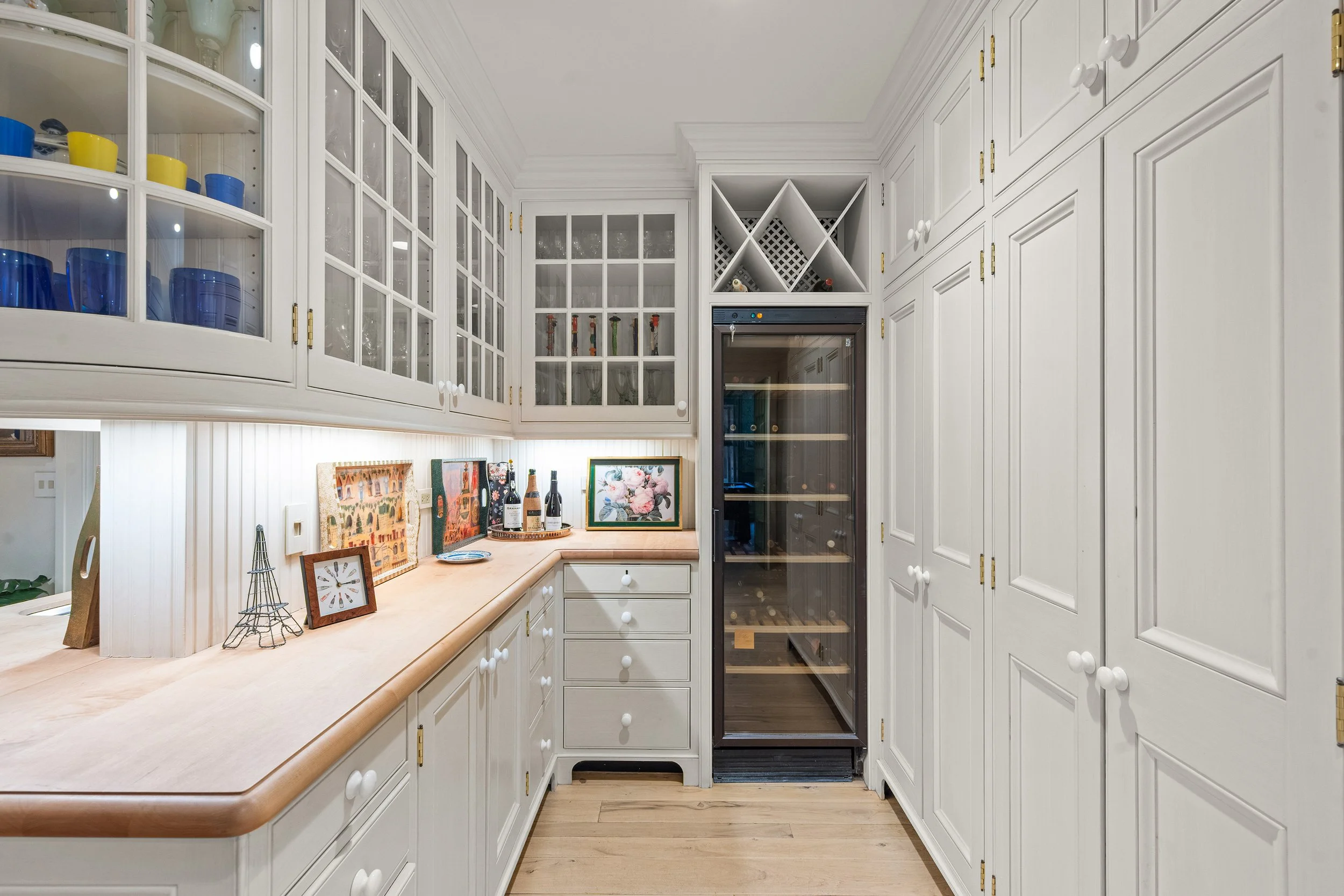 A kitchen corner with white cabinets, glass-fronted upper cabinets, a built-in wine fridge, and a wooden countertop decorated with framed pictures, bottles, and a miniature Eiffel Tower sculpture.