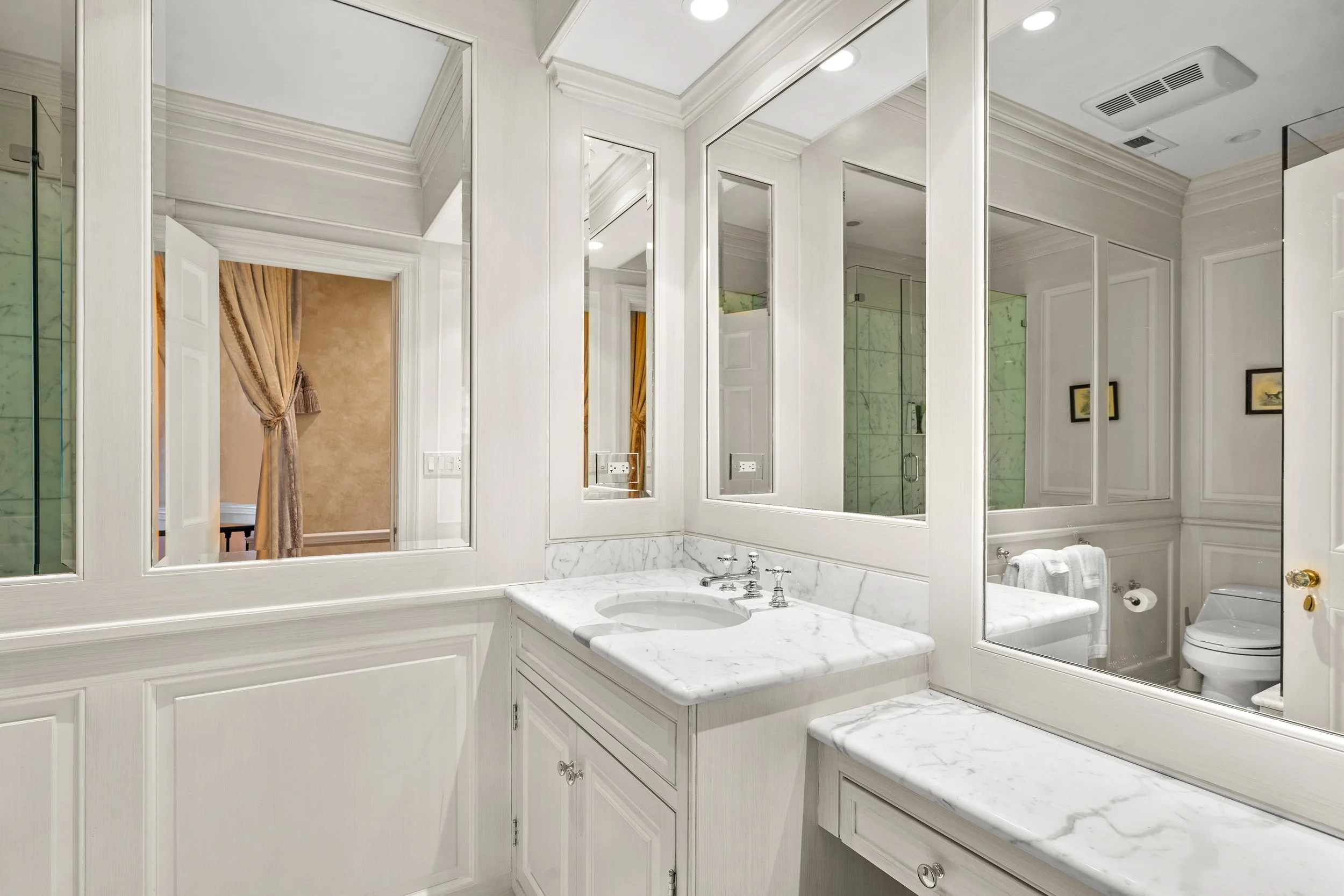 Bathroom with white cabinetry, marble countertops, and mirrors. Visible shower with glass doors and marble walls. Part of a toilet is visible through the mirror reflection.