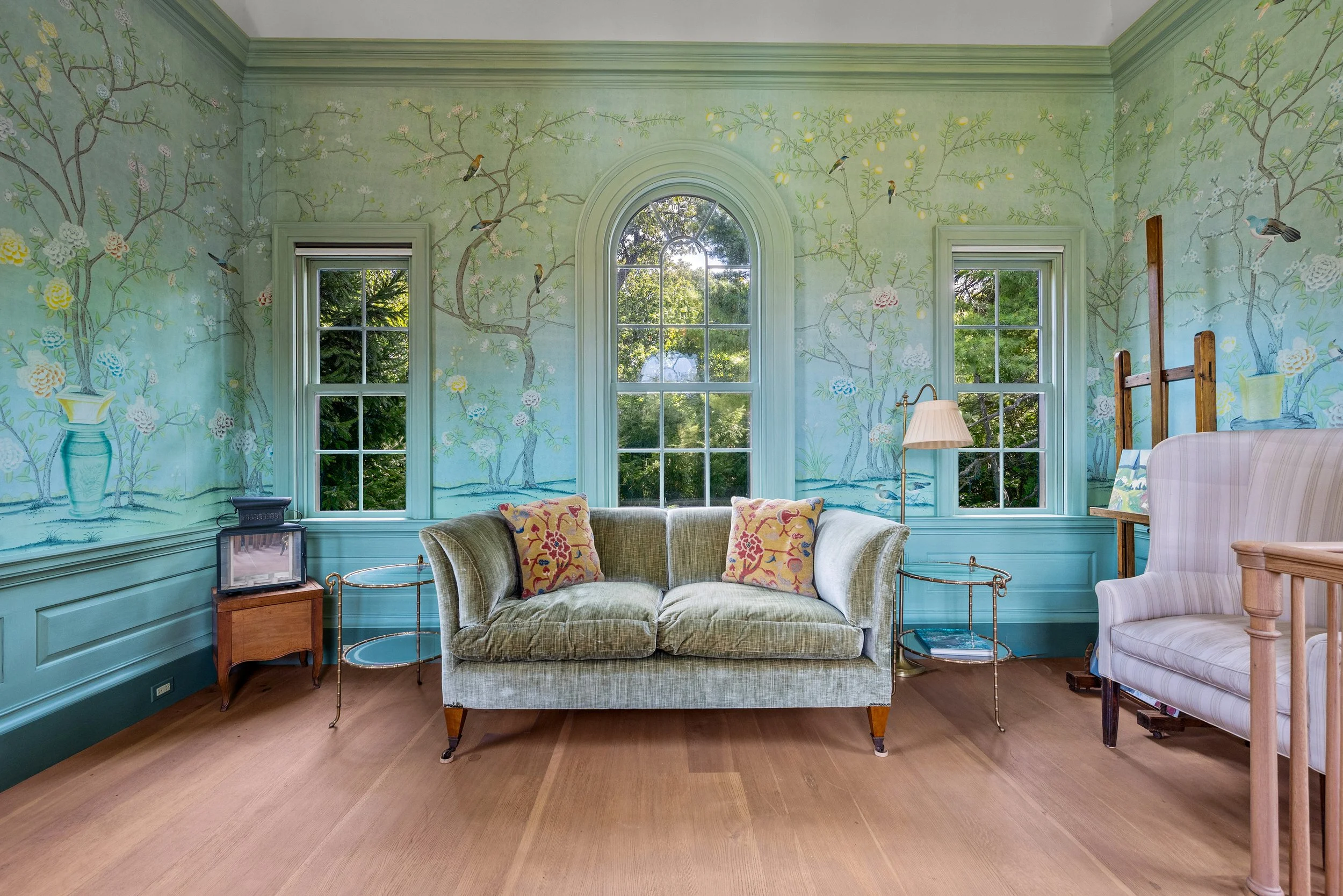 Living room with pastel green walls featuring painted trees and birds, three windows with greenery outside, olive green velvet sofa with two floral cushions, wooden side table with a small TV, floor lamp, and an armchair with a striped pattern, all on a wooden floor.