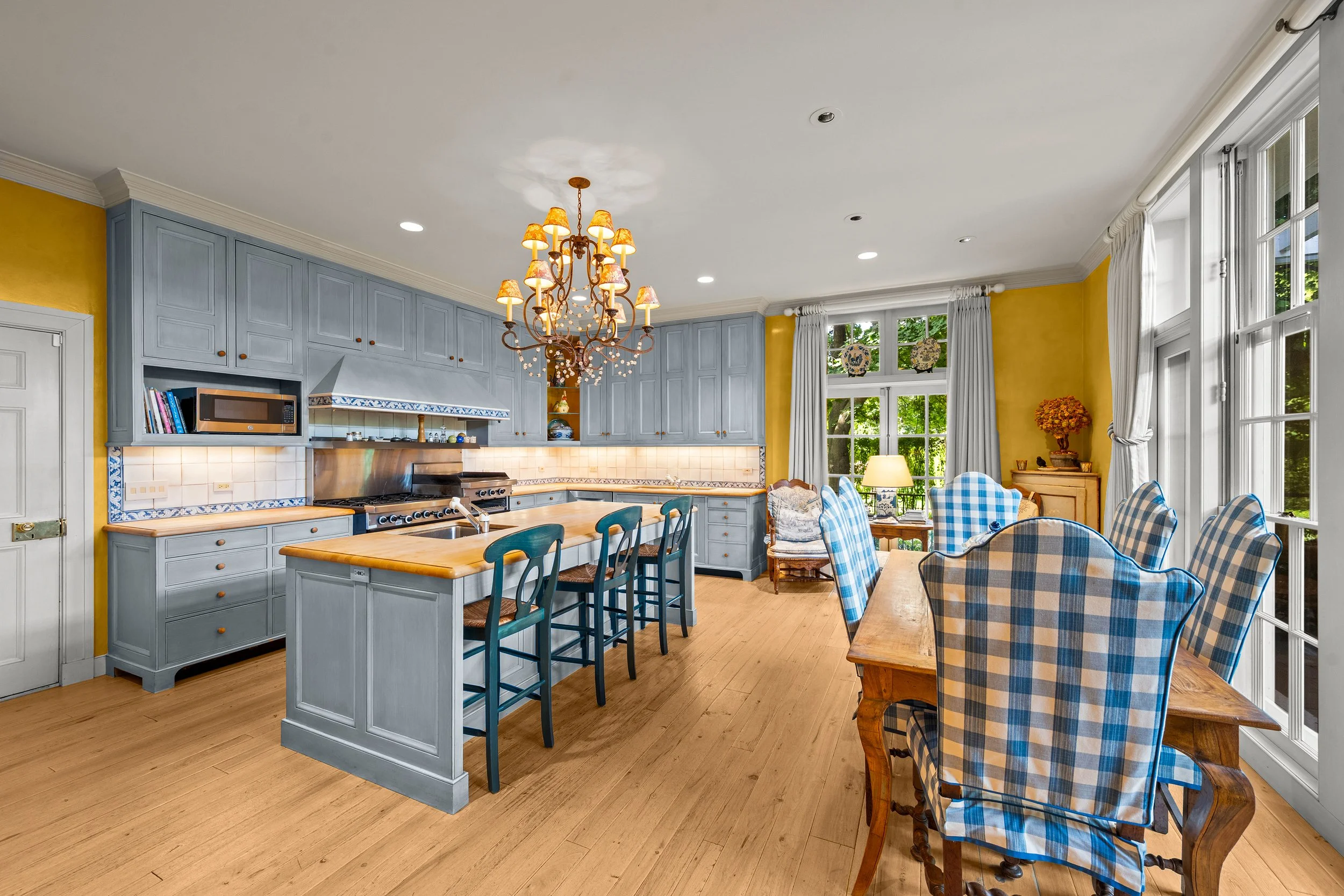 Kitchen and dining area with blue cabinetry, wooden island, plaid chairs, chandelier, and large windows with curtains, in a bright and cozy home.