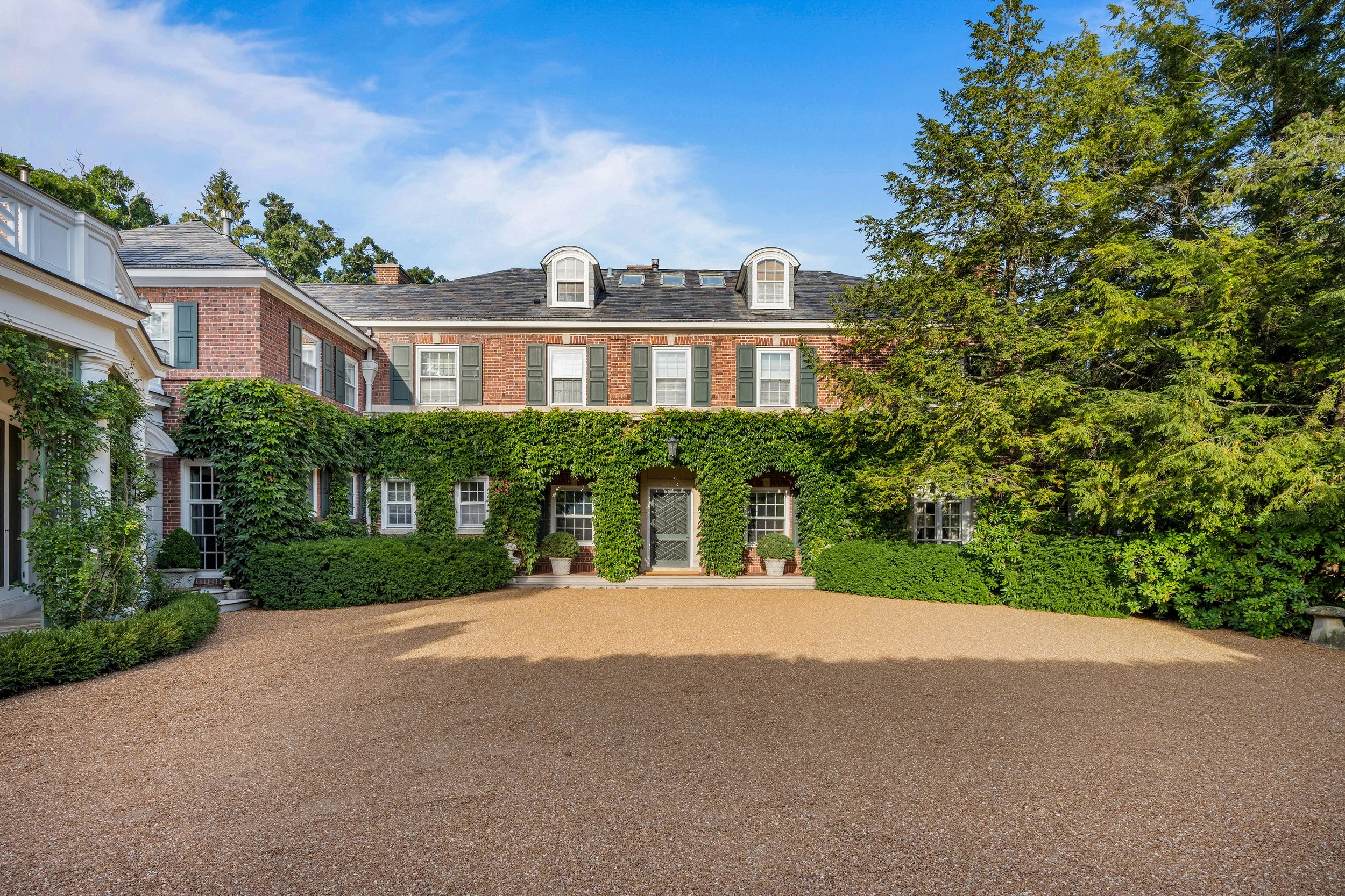 A large brick house with green shutters and ivy growing on the facade, surrounded by trees and lush greenery.