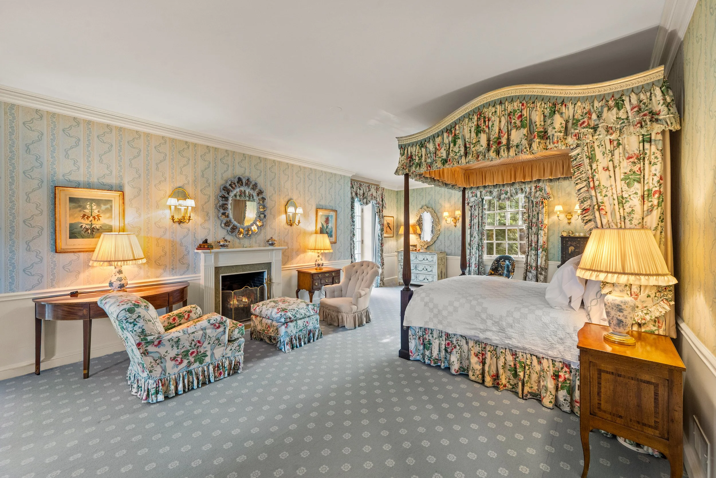 A vintage-style bedroom with floral drapes, a four-poster bed, and antique furniture, including lamps, chairs, and a fireplace.