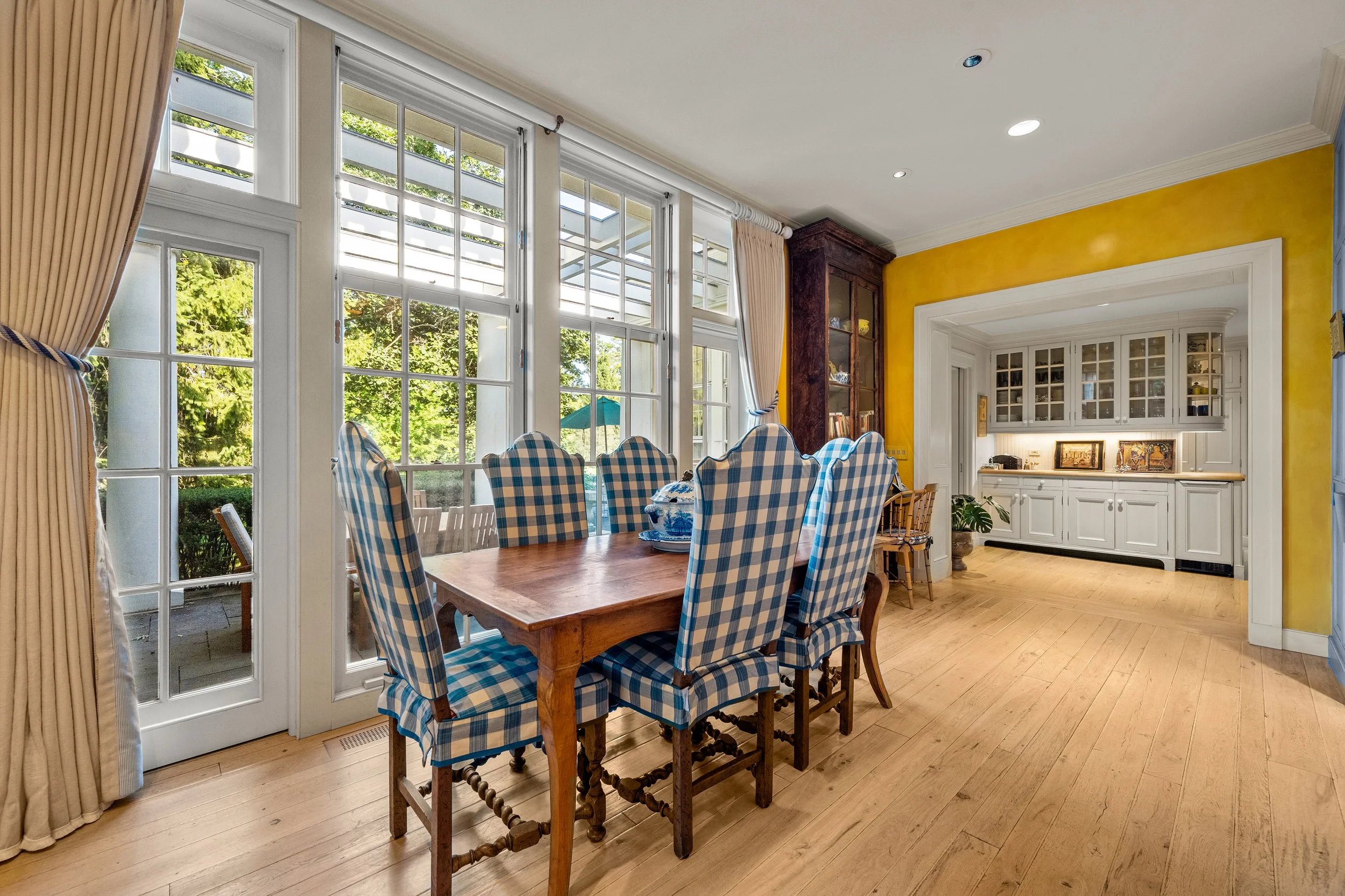 Dining room with a wooden table and checkered blue and white upholstered chairs, large windows with cream curtains, a built-in white cabinet, a yellow wall, and hardwood floors.