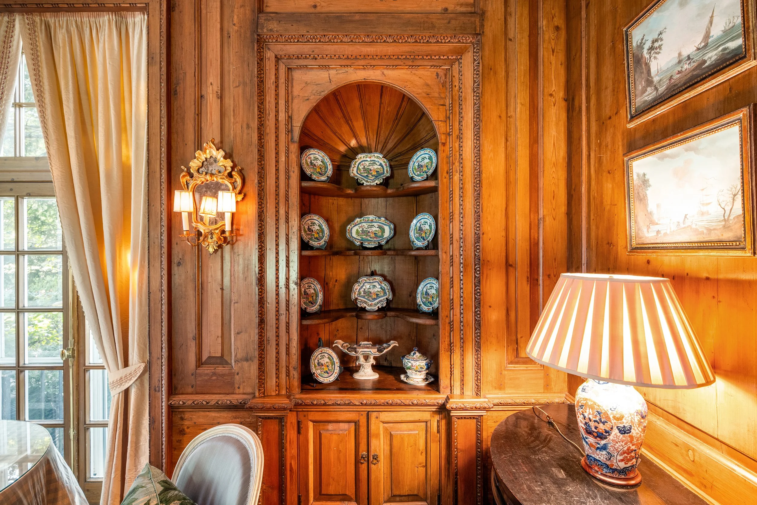 A wooden-paneled room with a built-in shelf displaying decorative porcelain plates and a bowl. To the left, a window with cream-colored curtains. On the right, a table with a floral ceramic lamp. Two framed landscape paintings are on the wooden wall.