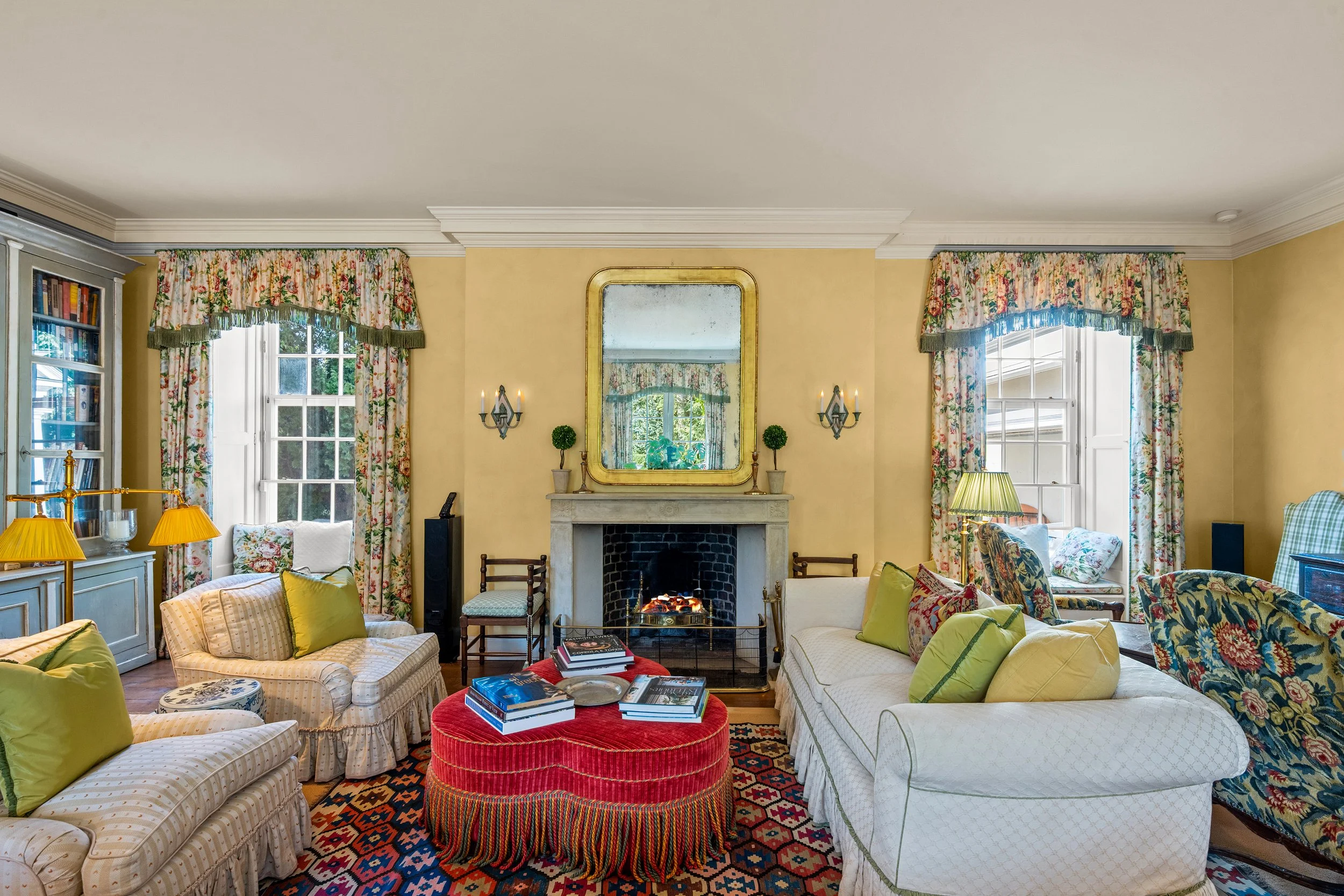 Living room with yellow walls, floral curtains, white sofa with green and yellow pillows, a fireplace with a mirror above it, and a patterned rug. There are chairs, lamps, books, and decorative items.