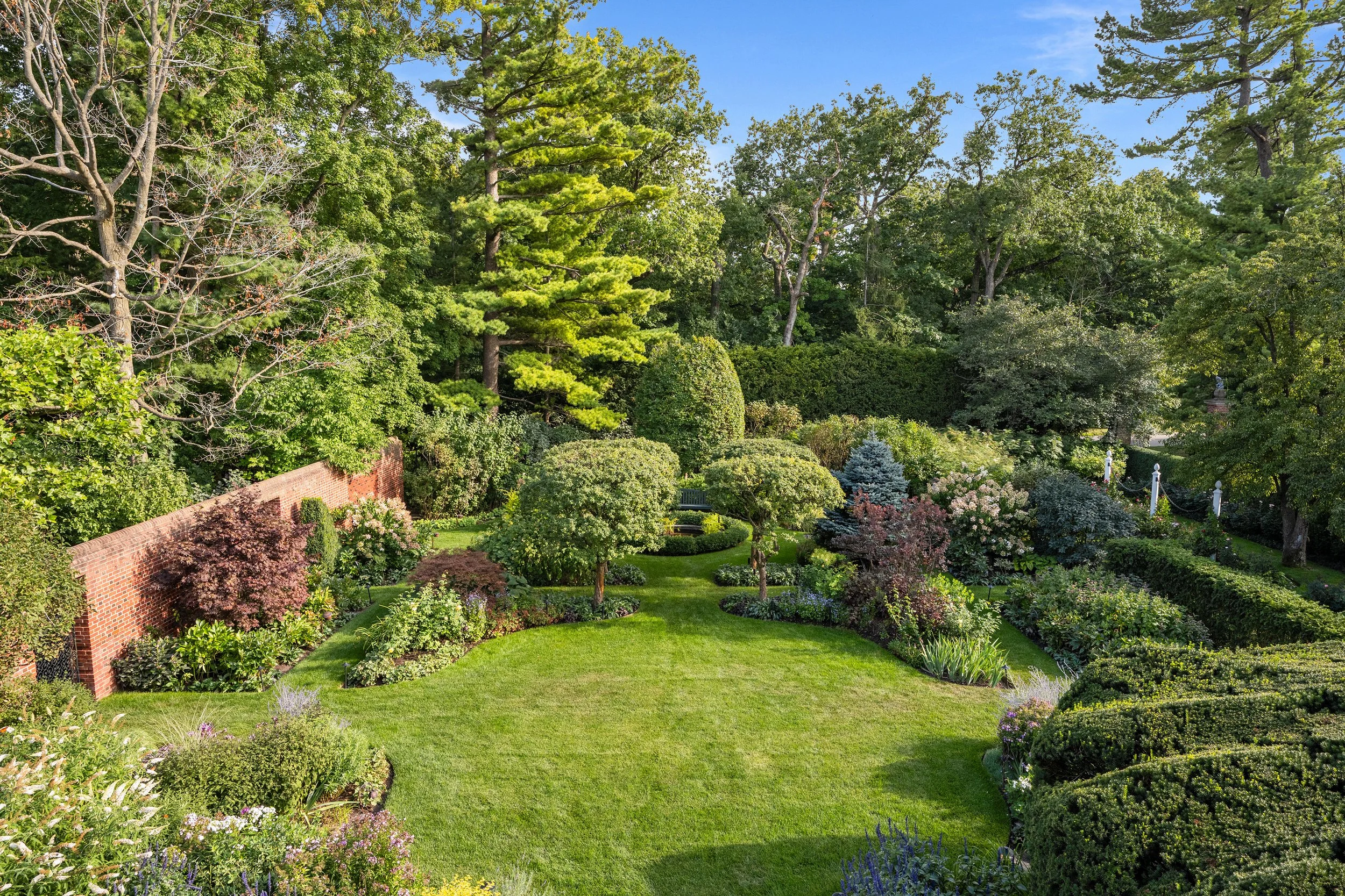 A well-maintained garden with a grassy lawn, trimmed bushes, and a variety of trees and flowering plants, bordered by a brick wall and surrounded by lush green foliage.