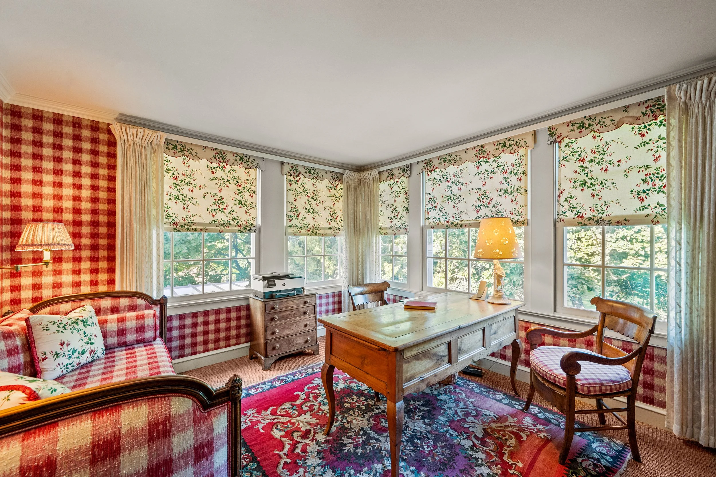 A cozy living room with red and green patterned wallpaper and matching window valances, a checkered sofa with floral cushions, a wooden desk with a lamp, a small dresser with a printer, and floral curtains, illuminated by natural light through multip