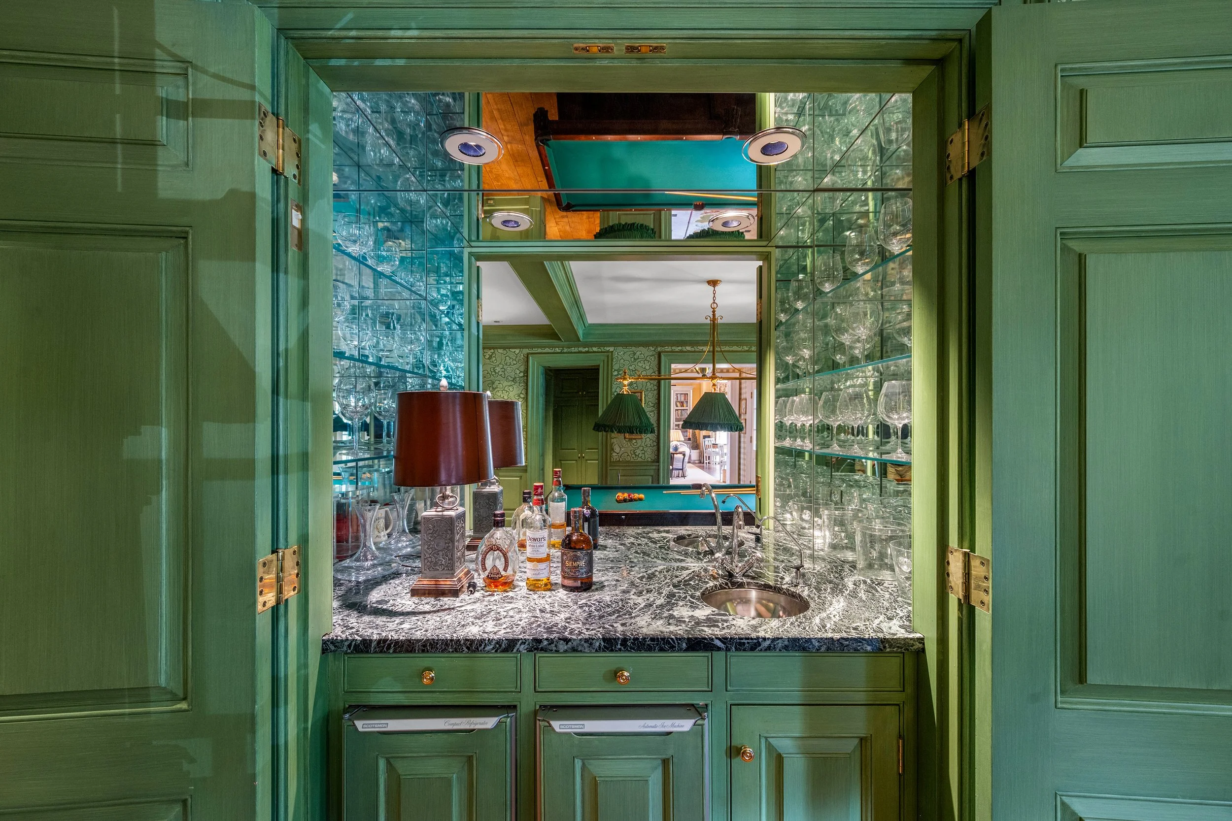 A view through an open green door revealing a bar area with a marble countertop, bottles of alcohol, a sink, table lamps, a mirror showing a room with green walls and a pool table, and glassware on shelves.