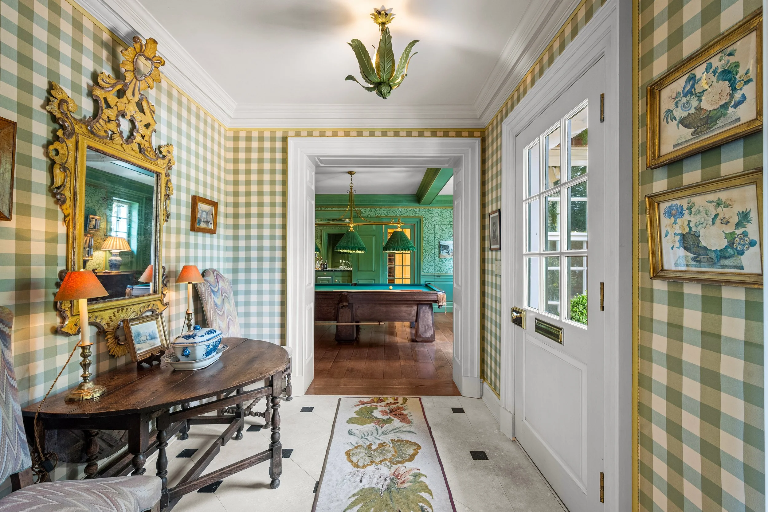 View of a dining room with green-checkered patterned wallpaper, gold framed artwork, and a wooden table with a lamp and decorative items. The room extends into another room with turquoise walls, a pool table, and hanging green pendant lights.