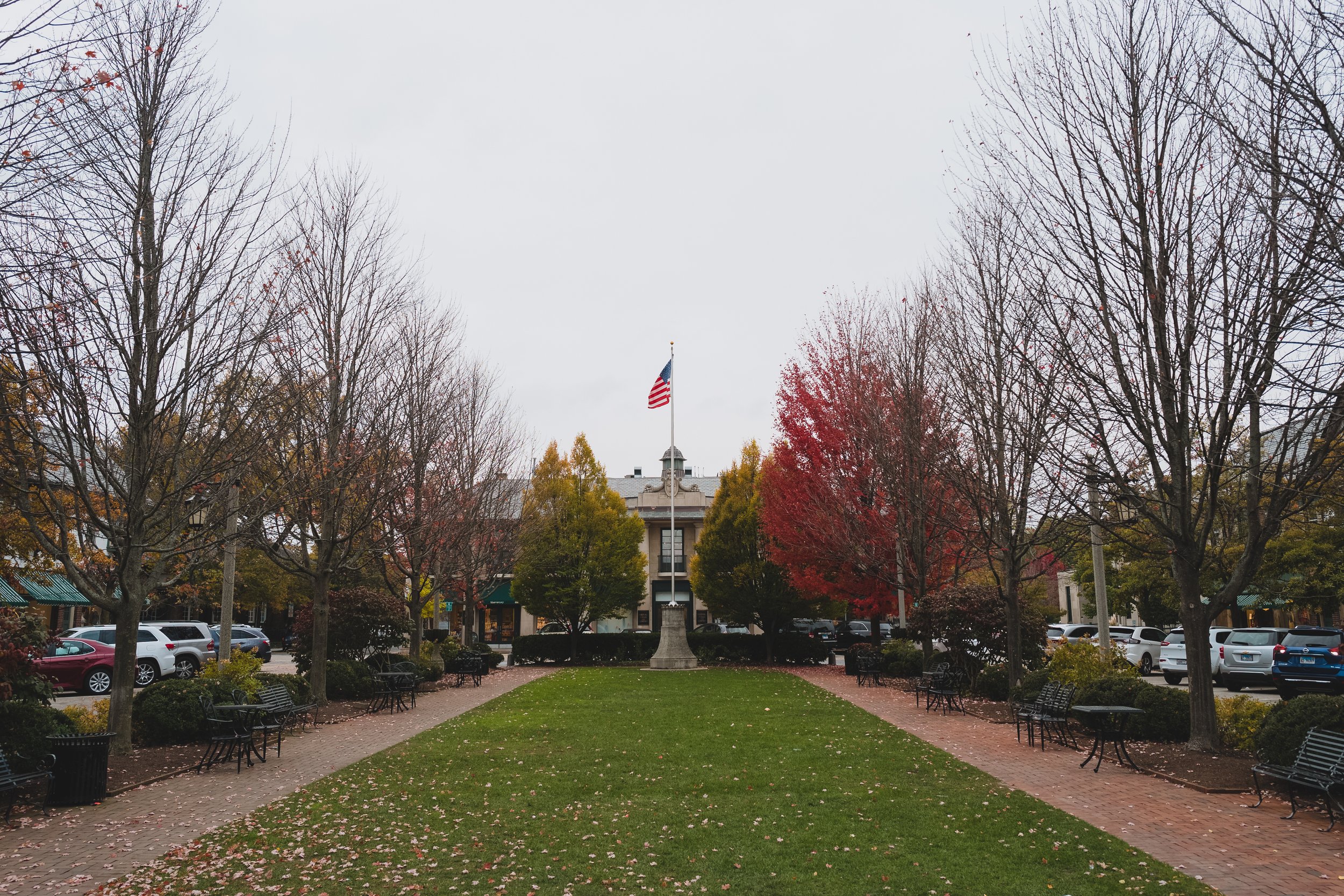 Lake Forest - A small park with a central flagpole flying the American flag, surrounded by trees with autumn-colored leaves and benches on brick pathways.