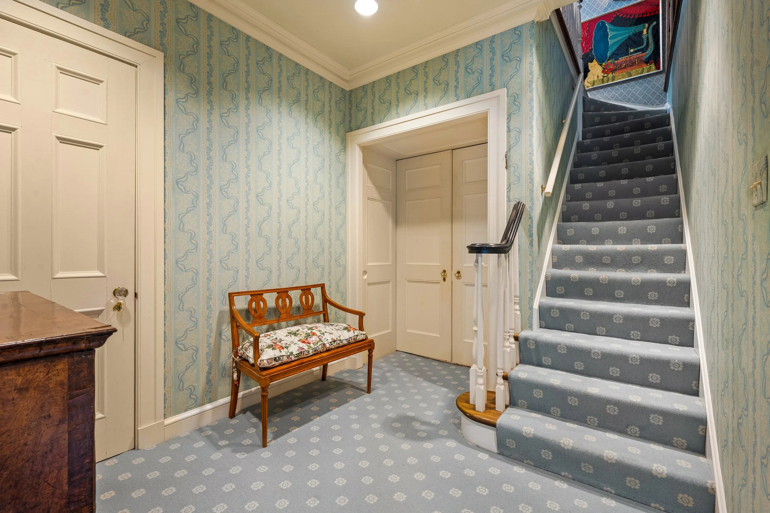 Interior of a vintage home with a staircase, bench, and double doors, decorated with patterned wallpaper and carpet.