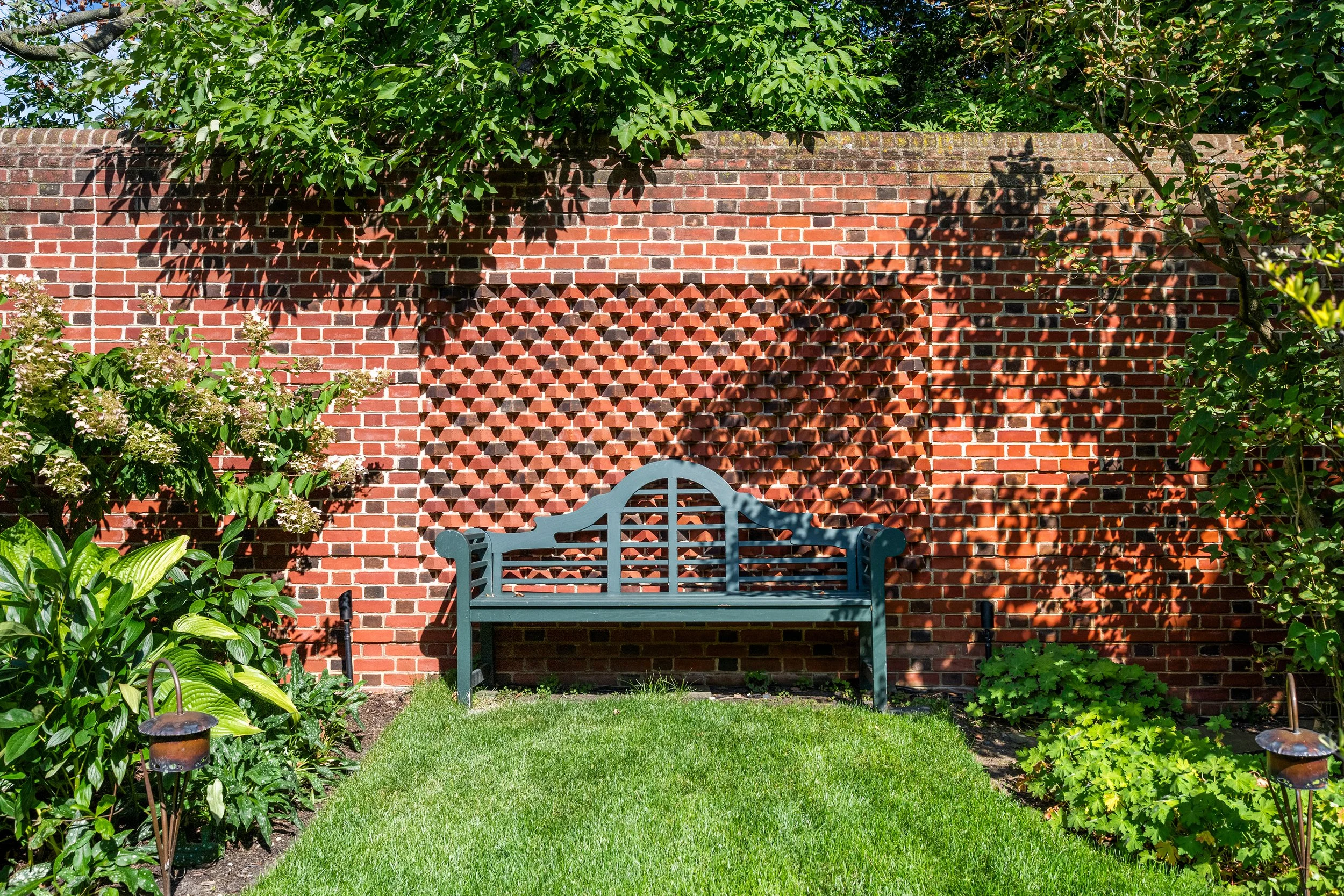 A garden scene with a blue bench in front of a brick wall. The wall has decorative brickwork patterns and shadows of tree branches and leaves cast on it. There are green plants and shrubs along the garden's perimeter, and sunlight illuminates the sce