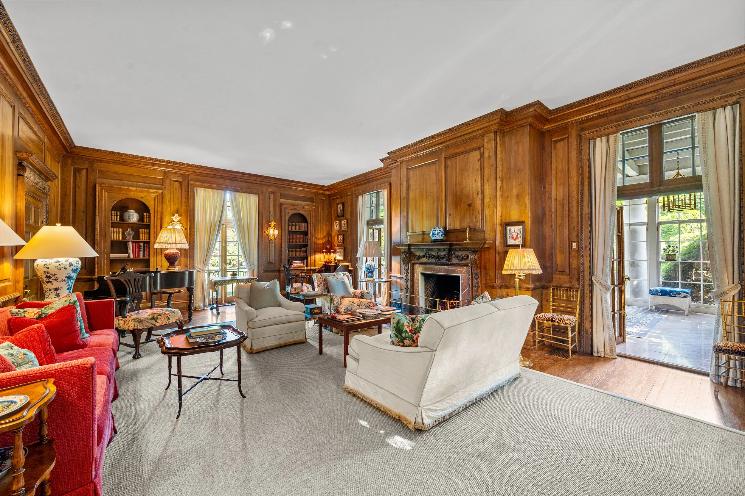 Living room with wood-paneled walls, white and floral sofas, colorful armchairs, a fireplace, a grand piano, and large windows with curtains.