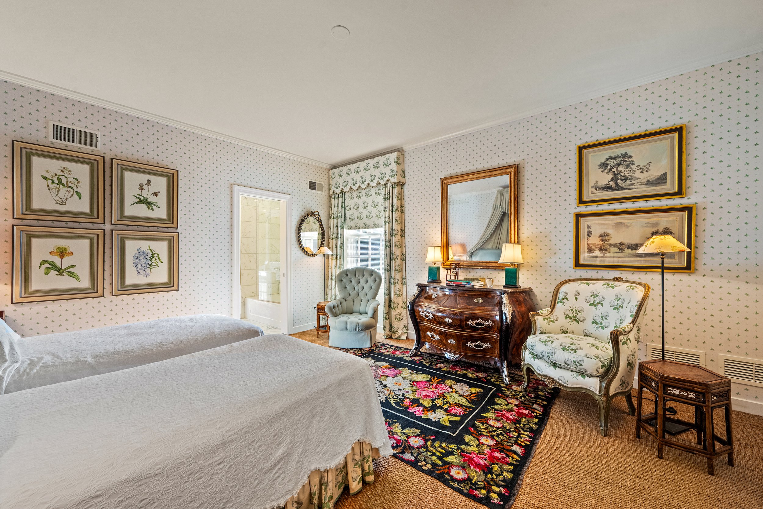 A vintage-style bedroom with two beds with white quilts, floral and botanical artwork on the wall, a floral-patterned window curtain, a floral area rug, and antique furniture including a wooden dresser, two upholstered armchairs, a mirror, and table 