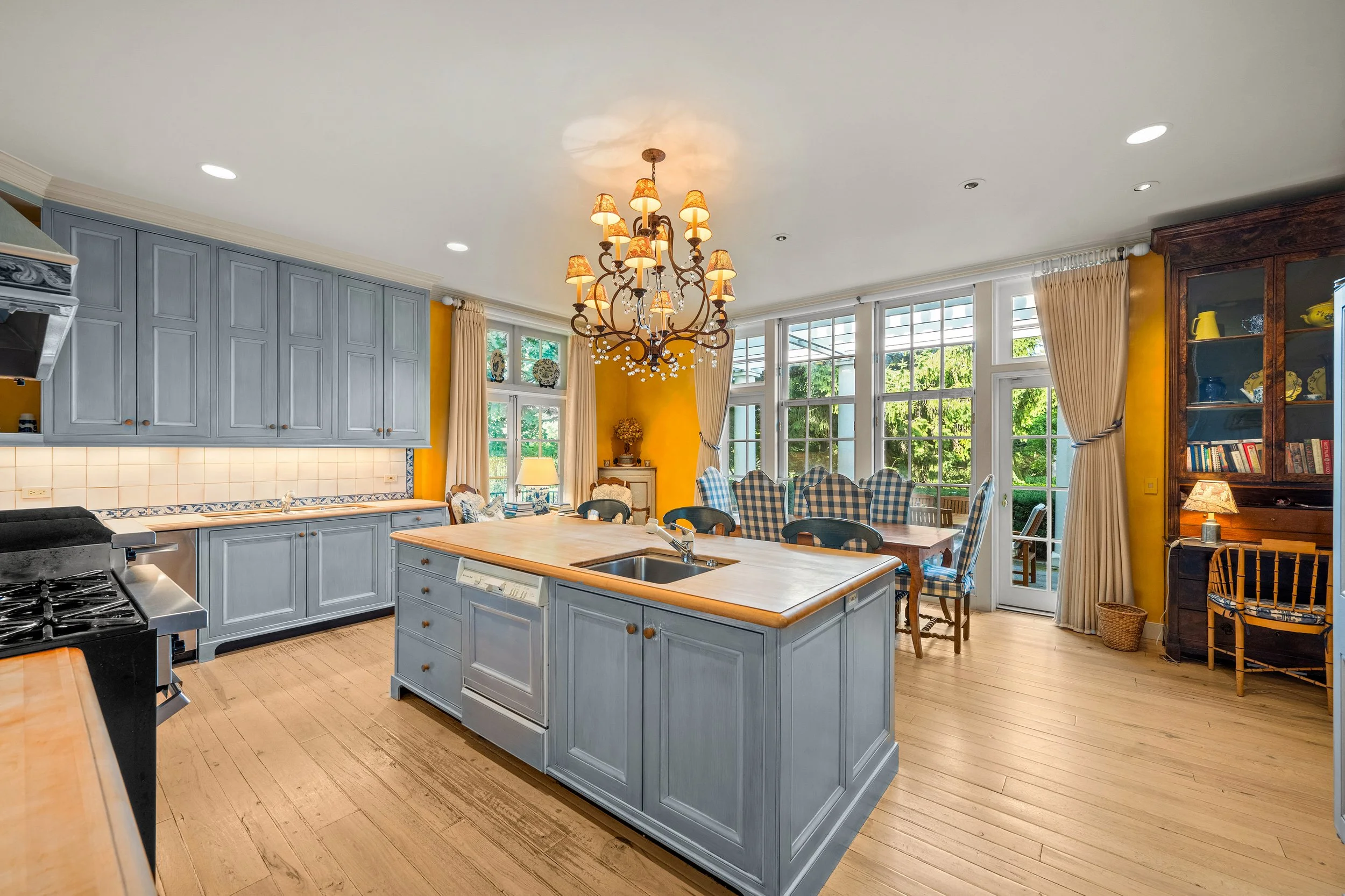 Bright kitchen with light blue cabinets, an island with a sink, a chandelier, large windows with cream curtains, and a dining area with checkered chairs.