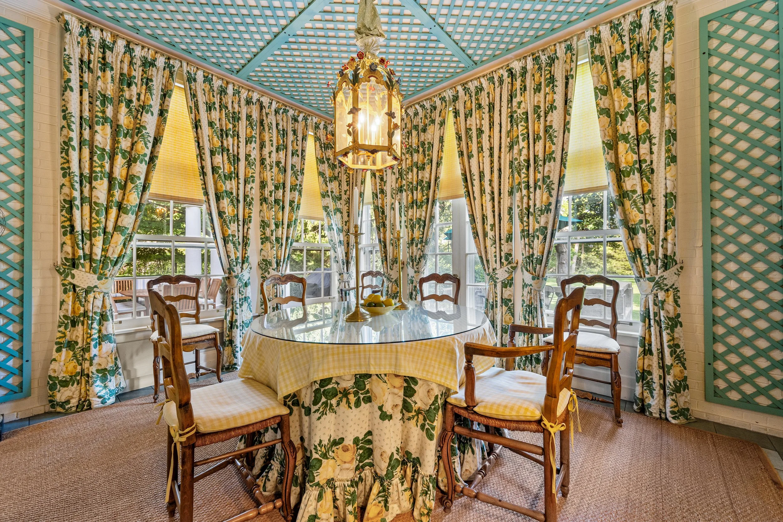 A dining room with a round glass table covered with a yellow and white checkered tablecloth, surrounded by five wooden chairs with upholstered seats. The room has large windows with floral curtains, a chandelier hanging from a lattice ceiling painted