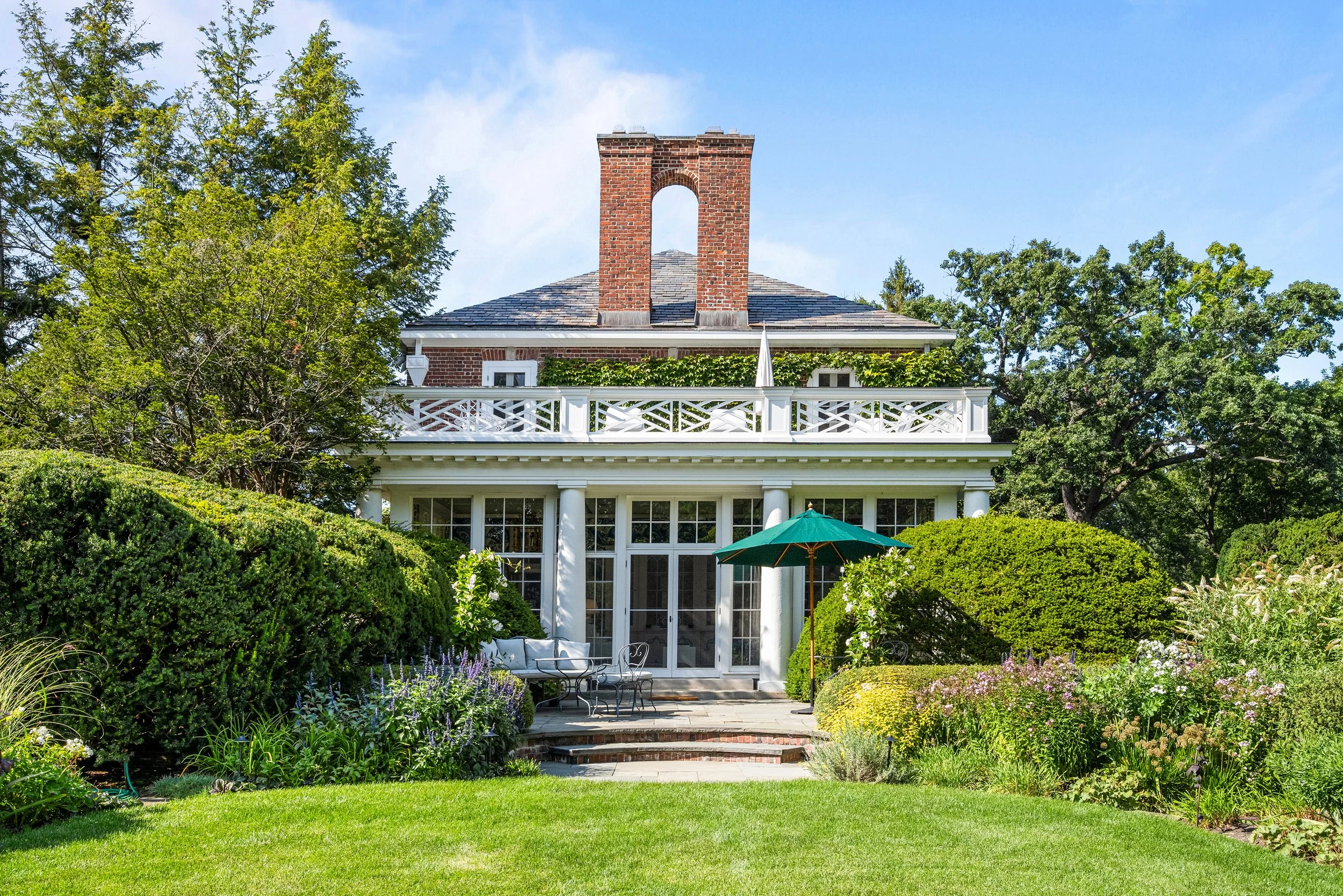 Backyard garden view of a two-story house with large glass doors, white porch railing, and a brick chimney, surrounded by lush green trees and flowering plants.