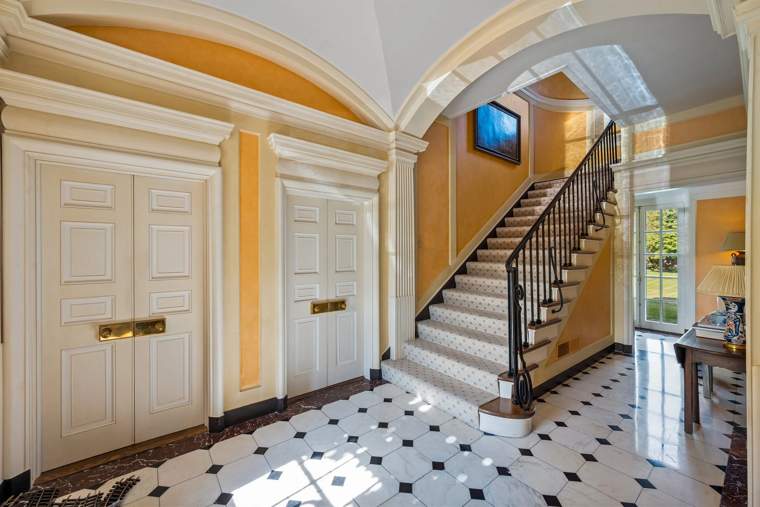 Elegant foyer with white double doors, a staircase with a carpet runner, marble tiled floor, and a side table with lamps near a glass door leading outside.