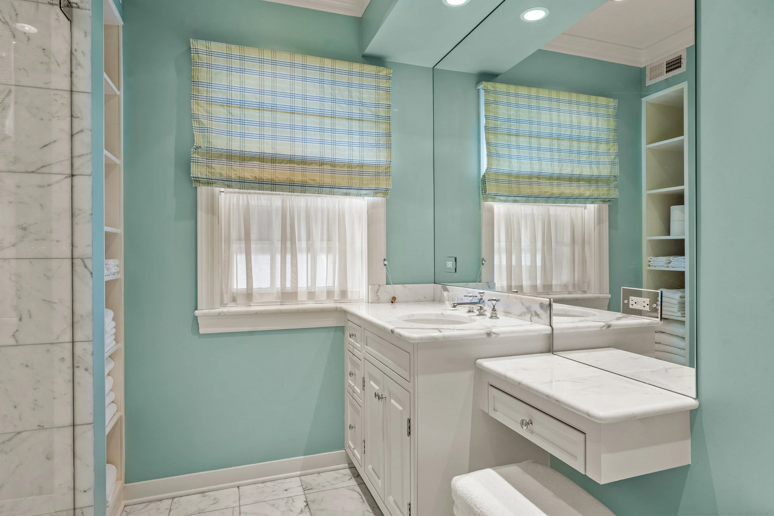 Bathroom with a large mirror, white marble sink, green walls, two windows with plaid and sheer curtains, and built-in shelves with towels.