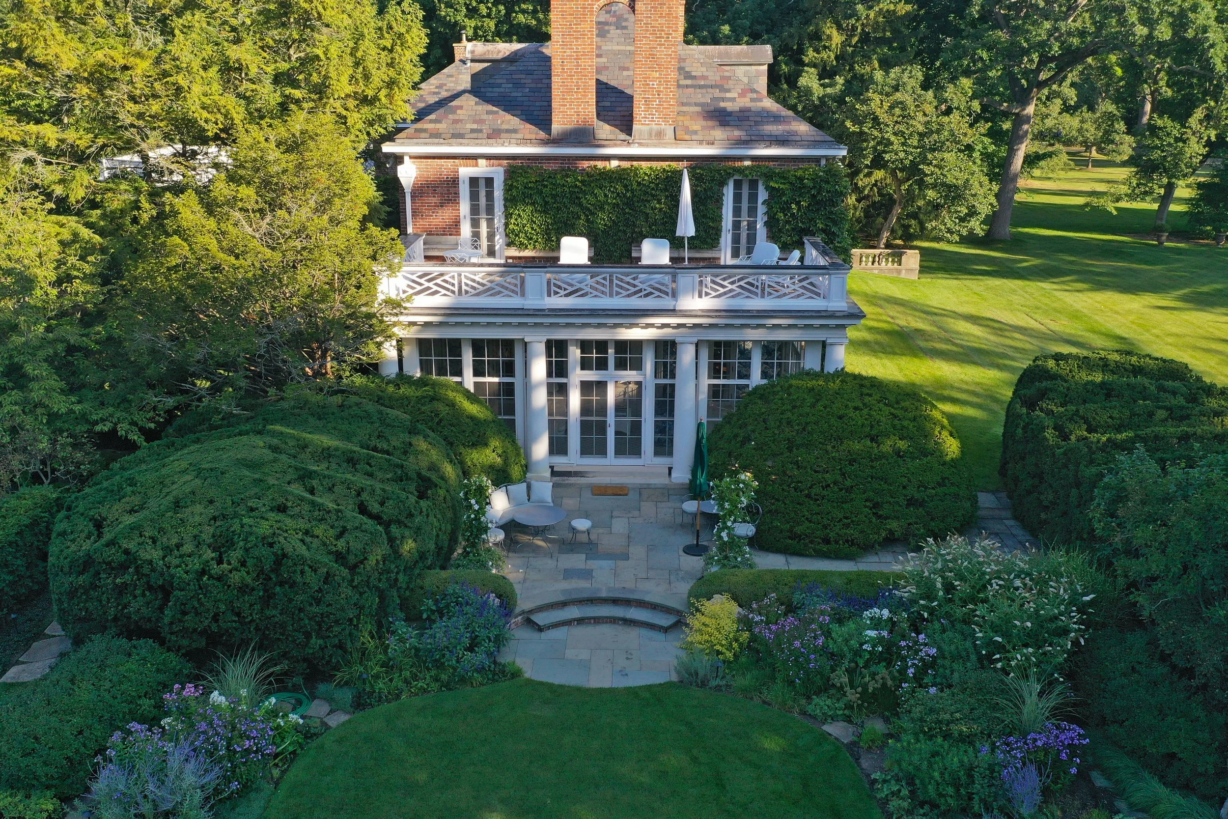 An aerial view of a two-story house with a brick exterior and a slate roof. The house features large glass windows and a spacious deck with outdoor furniture, including chairs, a table, and a large umbrella. The backyard includes lush green grass, ne