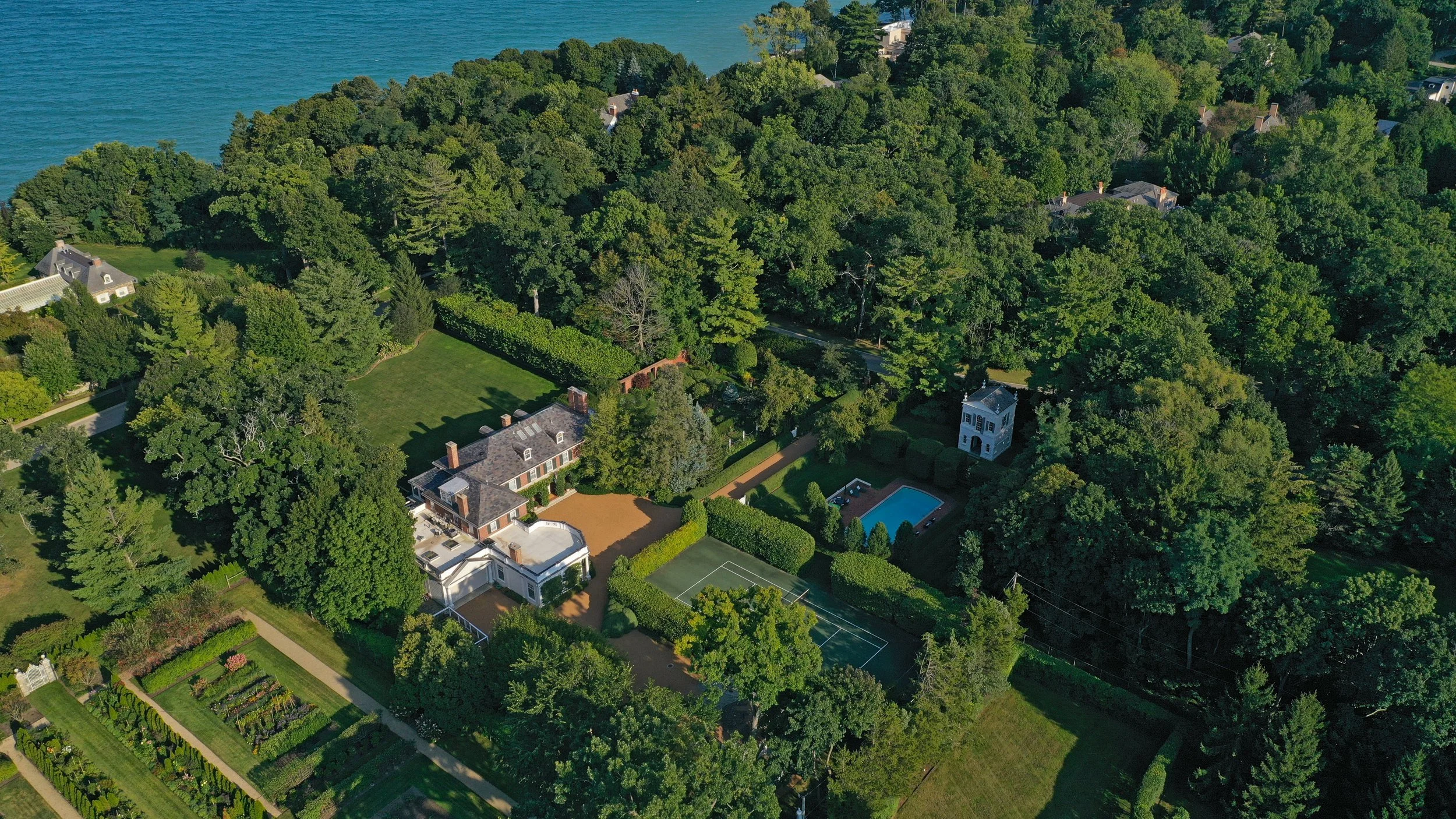 Aerial view of a large estate with a historic house, tennis courts, swimming pool, tennis courts, garden, and dense trees near a body of water.