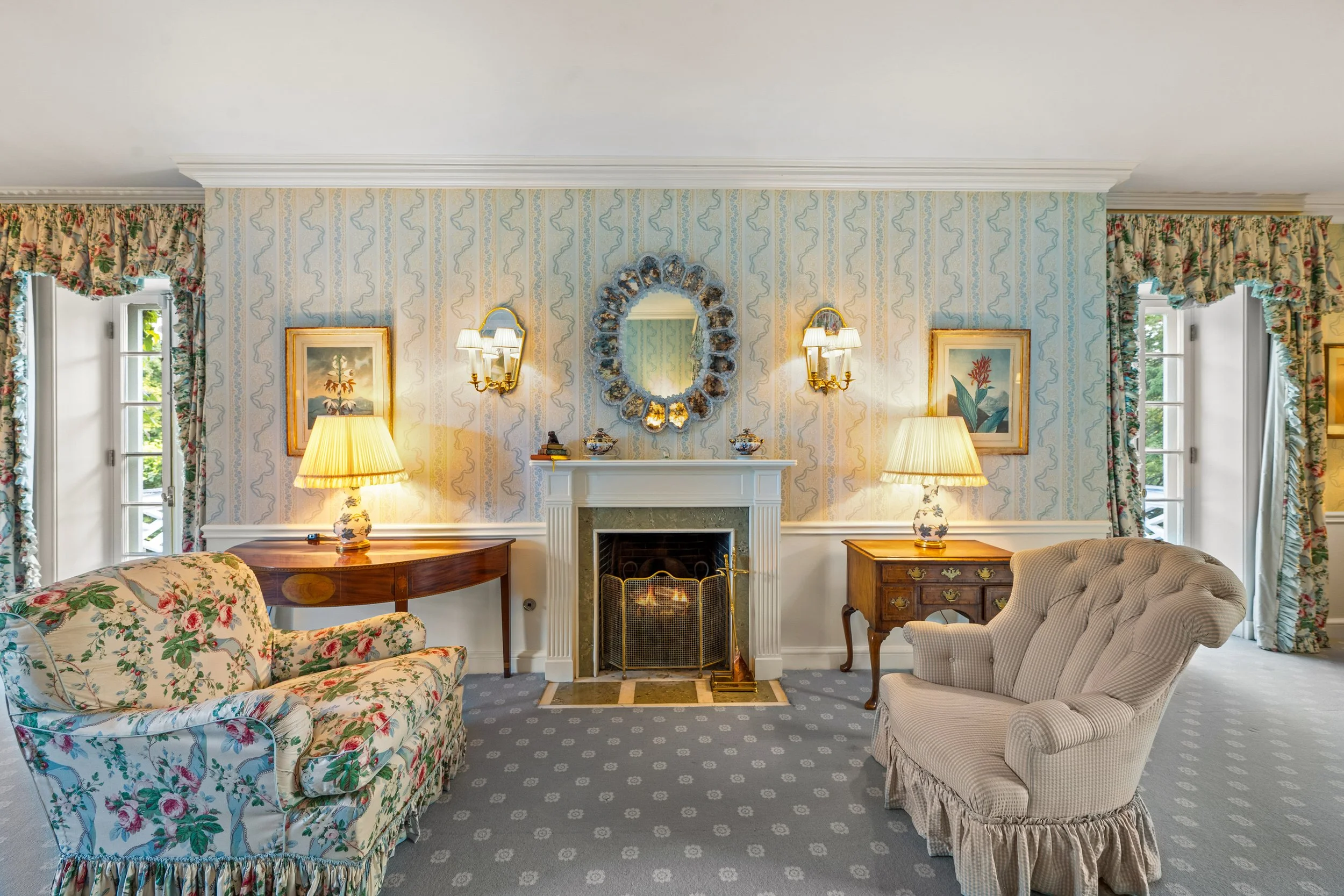 Living room with floral wallpaper, two windows with floral curtains, fireplace with mirror above, two table lamps, floral sofa on the left, beige armchair on the right.