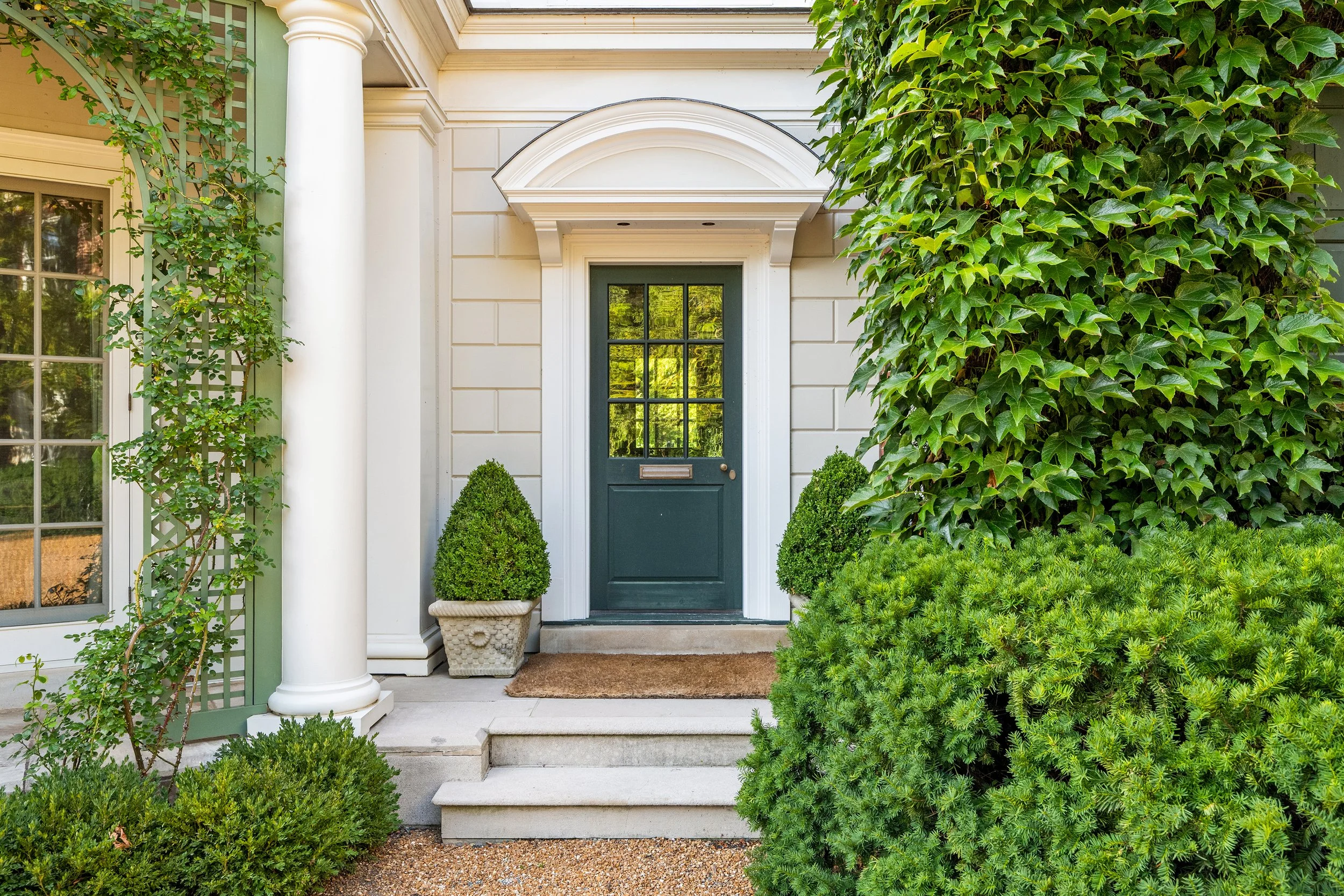Front door of a house with a dark green door, surrounded by lush green bushes and plants, with steps leading up to the door and white framing.